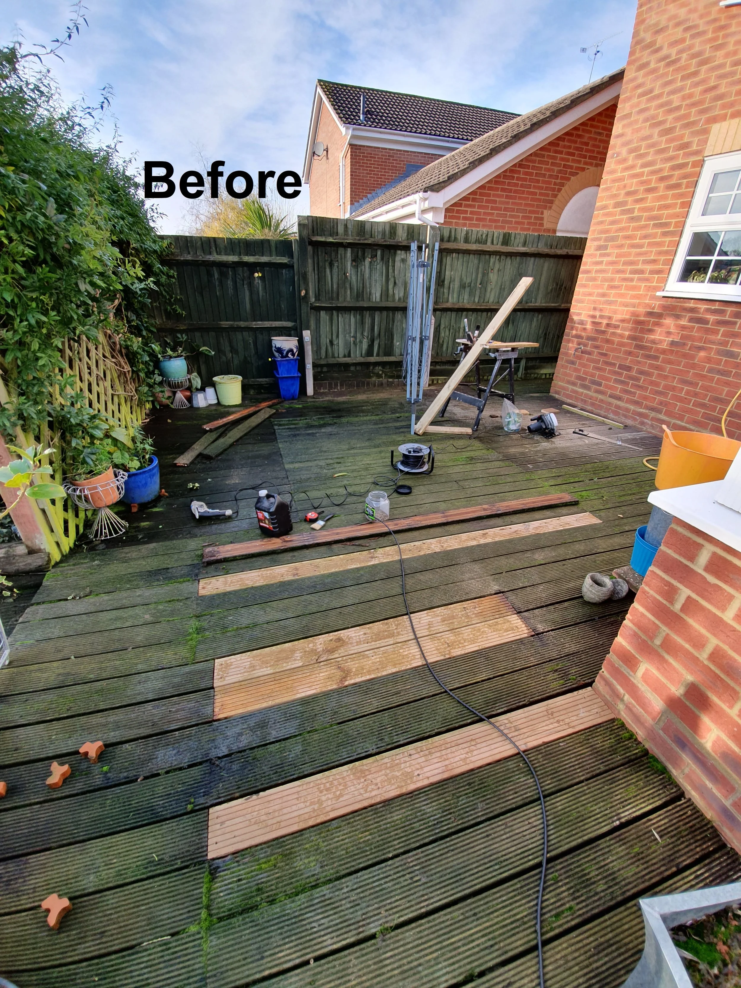 Backyard patio under renovation with wooden planks, tools, and equipment scattered around. The deck is partially replaced with new wood pieces awaiting installation.
