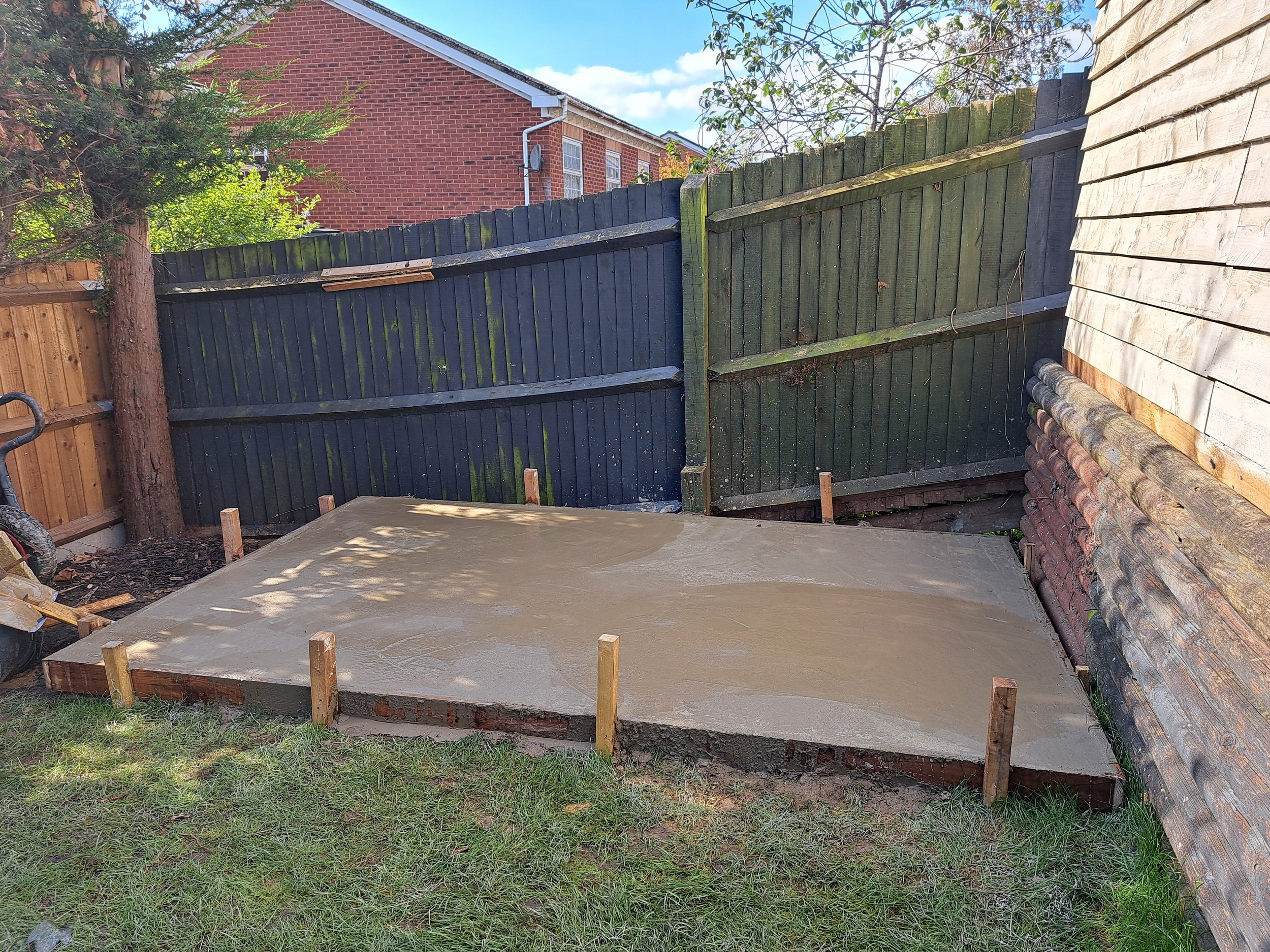 A small backyard patio with freshly poured concrete, surrounded by wooden fence panels, some of which are painted dark green and others unpainted, with a brick wall on the right side and a tree on the left.