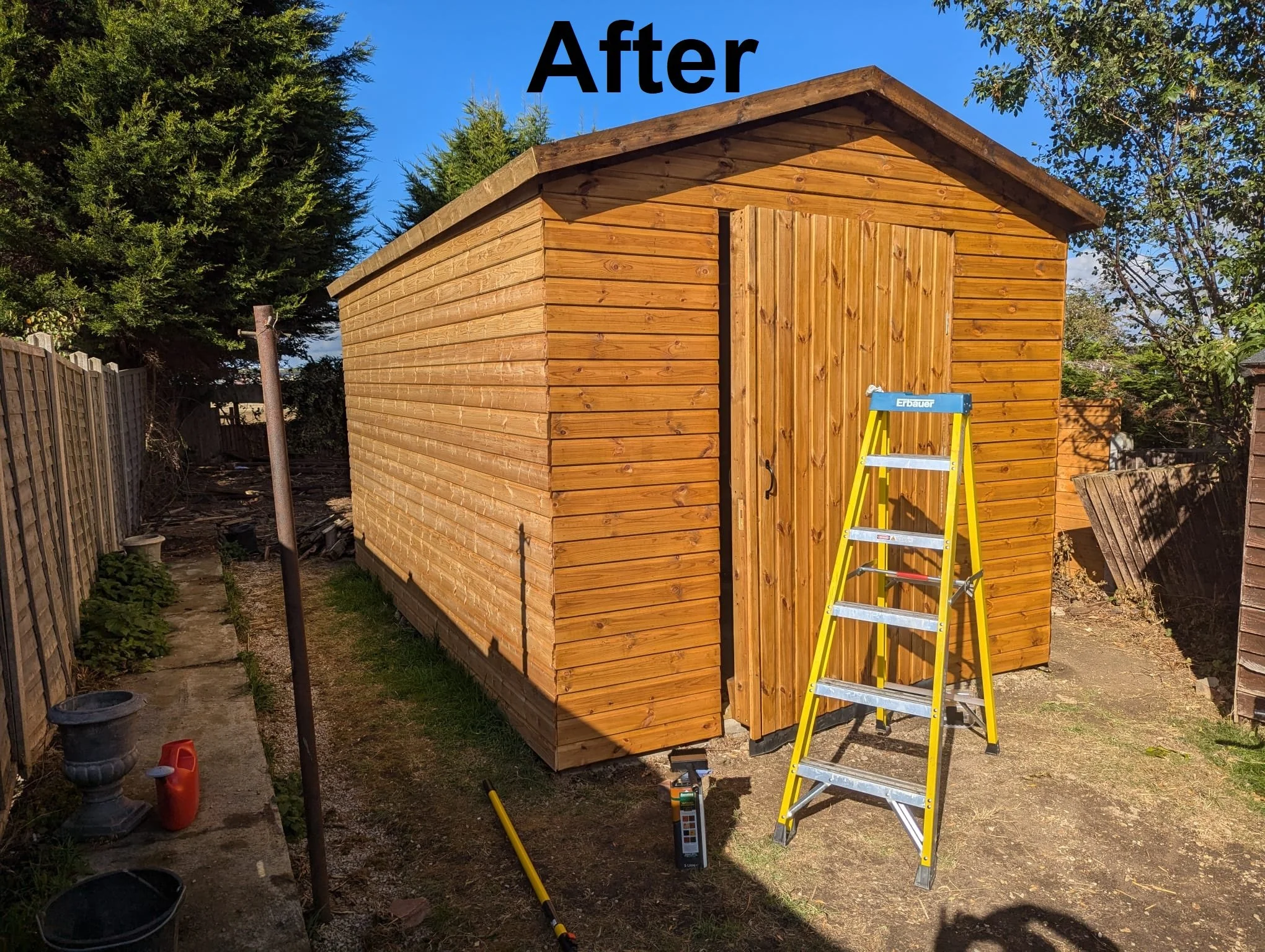 A newly built wooden shed with a steep gable roof, standing on a patch of grass and dirt. There is a yellow step ladder leaning against the shed, and various tools and supplies are on the ground nearby. Tall trees and a fence are in the background. T