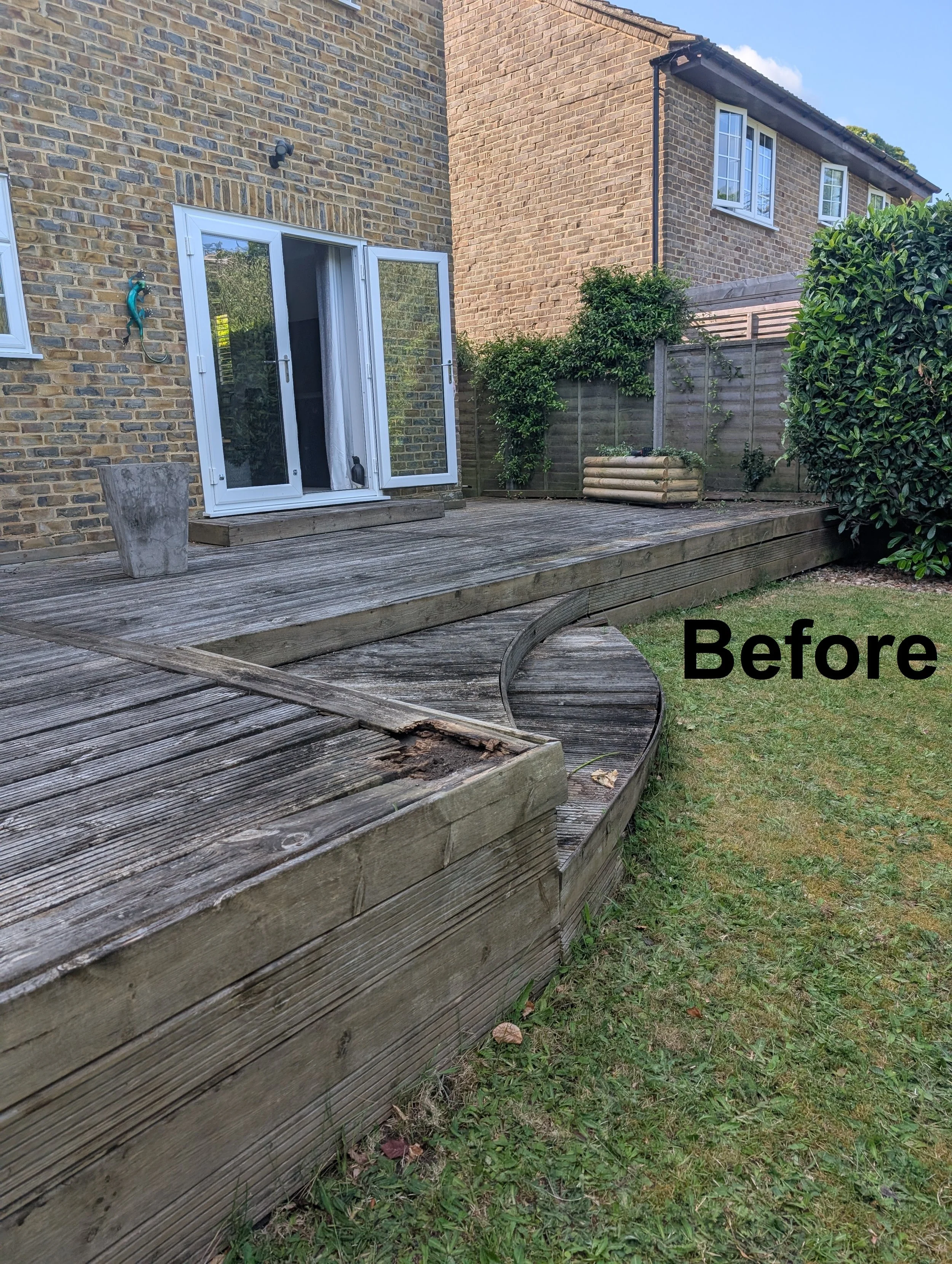 Backyard with wooden deck and grassy lawn, brick house with open French doors, and neighboring house in the background. The deck has some damage, with a section broken and warped.