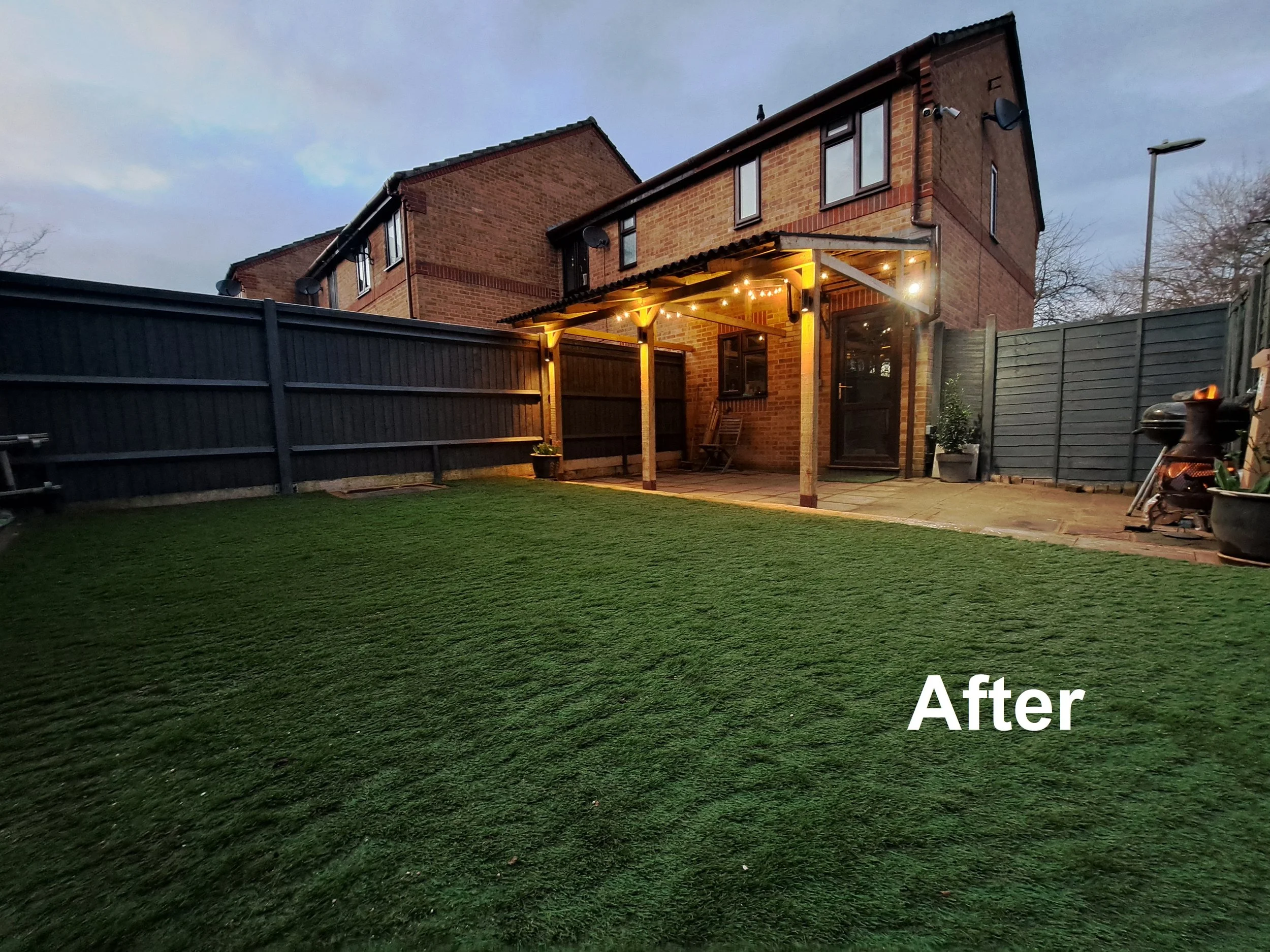Backyard with a lush green lawn and a new wooden patio cover with string lights, attached to a brick house with a black door and windows. There is a dark grey fence surrounding the yard, some potted plants, and garden equipment.