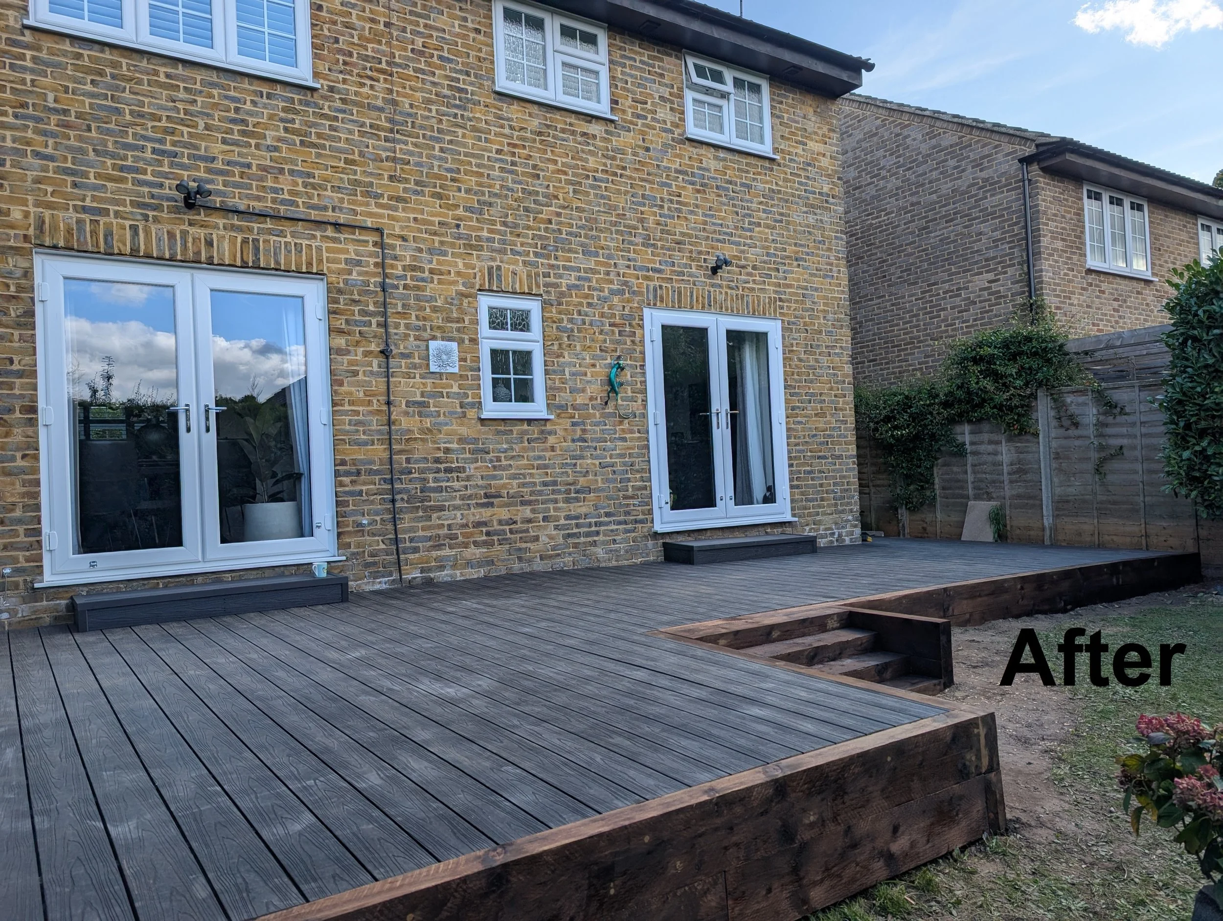 A backyard with a freshly built wooden deck attached to a brick house, with sliding glass doors and windows. There are stairs leading down from the deck, and some plants and trees along the fence line. The sky is partly cloudy.