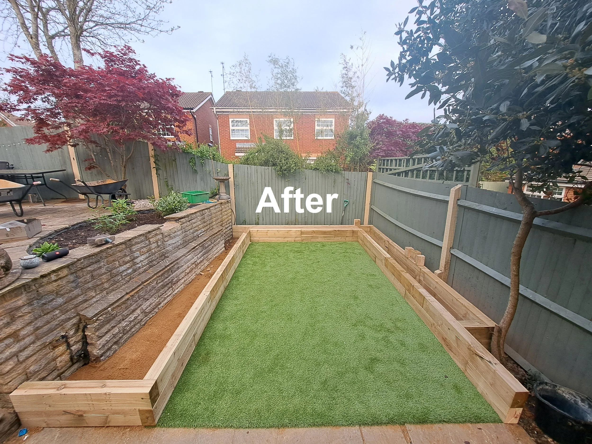 Backyard garden with new wooden frame, artificial grass, and surrounding trees and fence, showing a completed landscaping project.