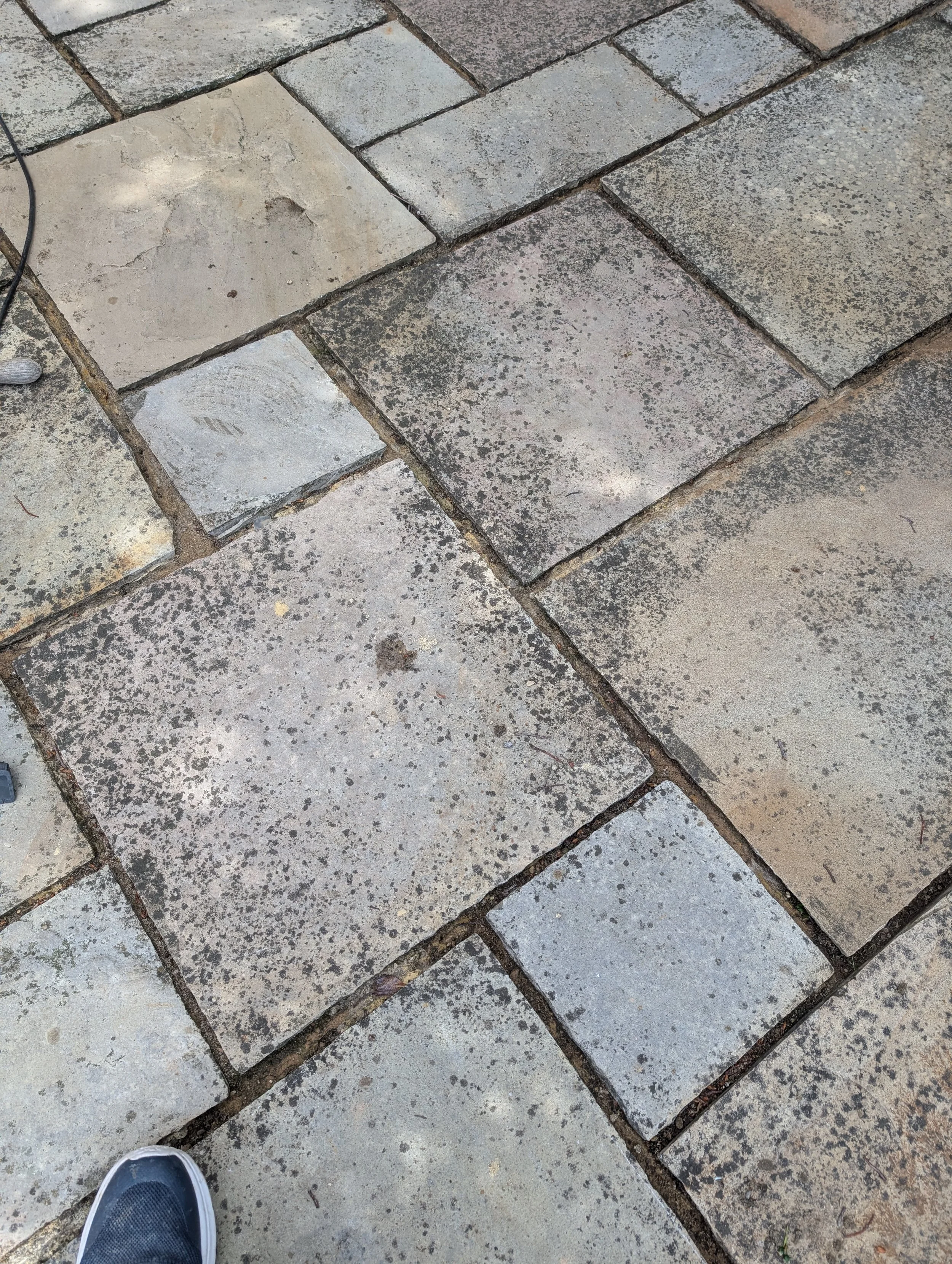 Close-up of a stone patio with rectangular light and dark gray stones with speckled textures, some with slight cracks, with a partial view of a person's foot in a gray and black sneaker at the bottom left corner.
