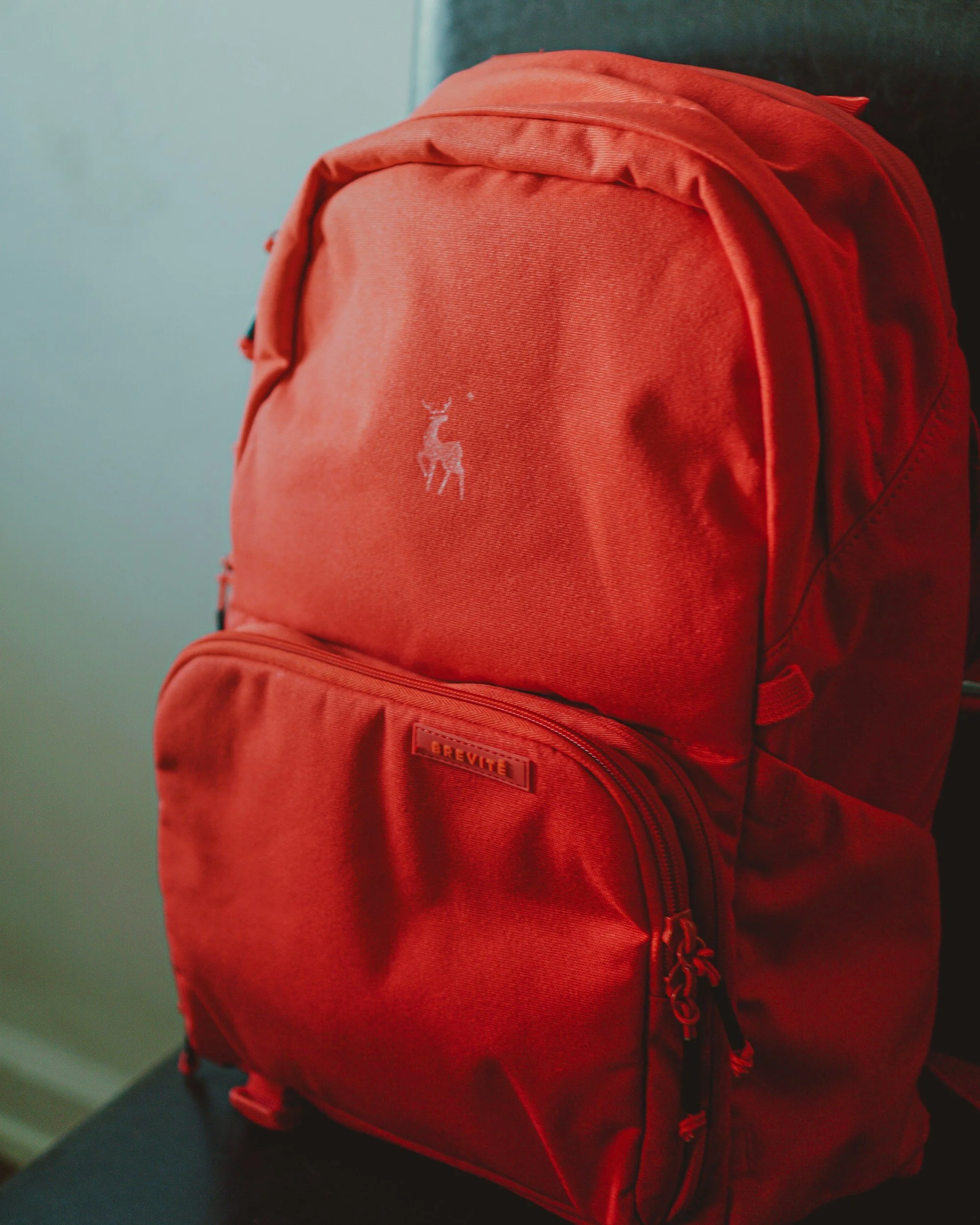A red backpack with a small logo of a deer on the upper front and a pocket with a zipper on the lower front, placed on a dark surface.