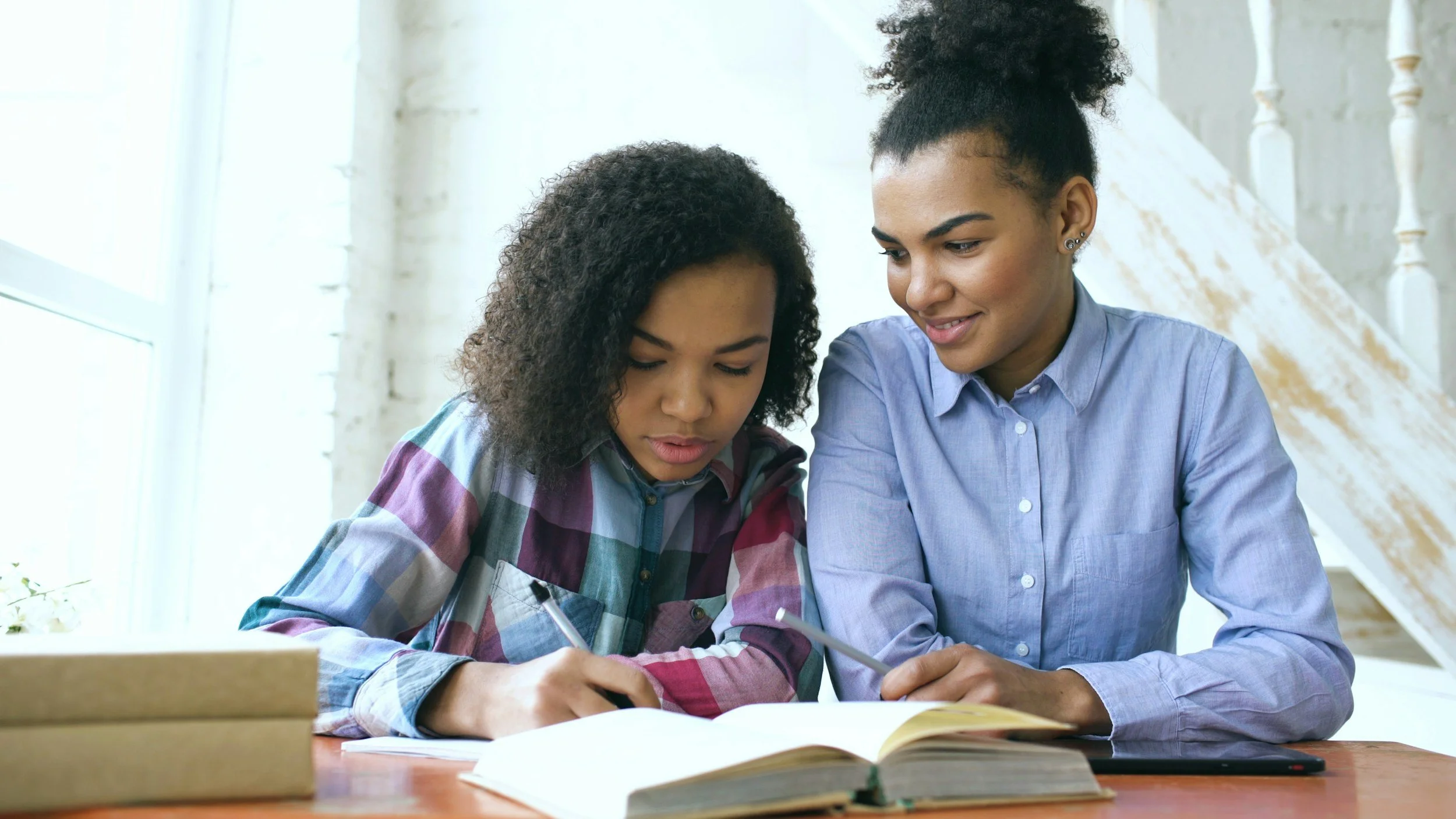 Two women sitting at a desk in a bright room, studying together with open books in front of them. One is writing in a notebook, the other is looking at the book and smiling.