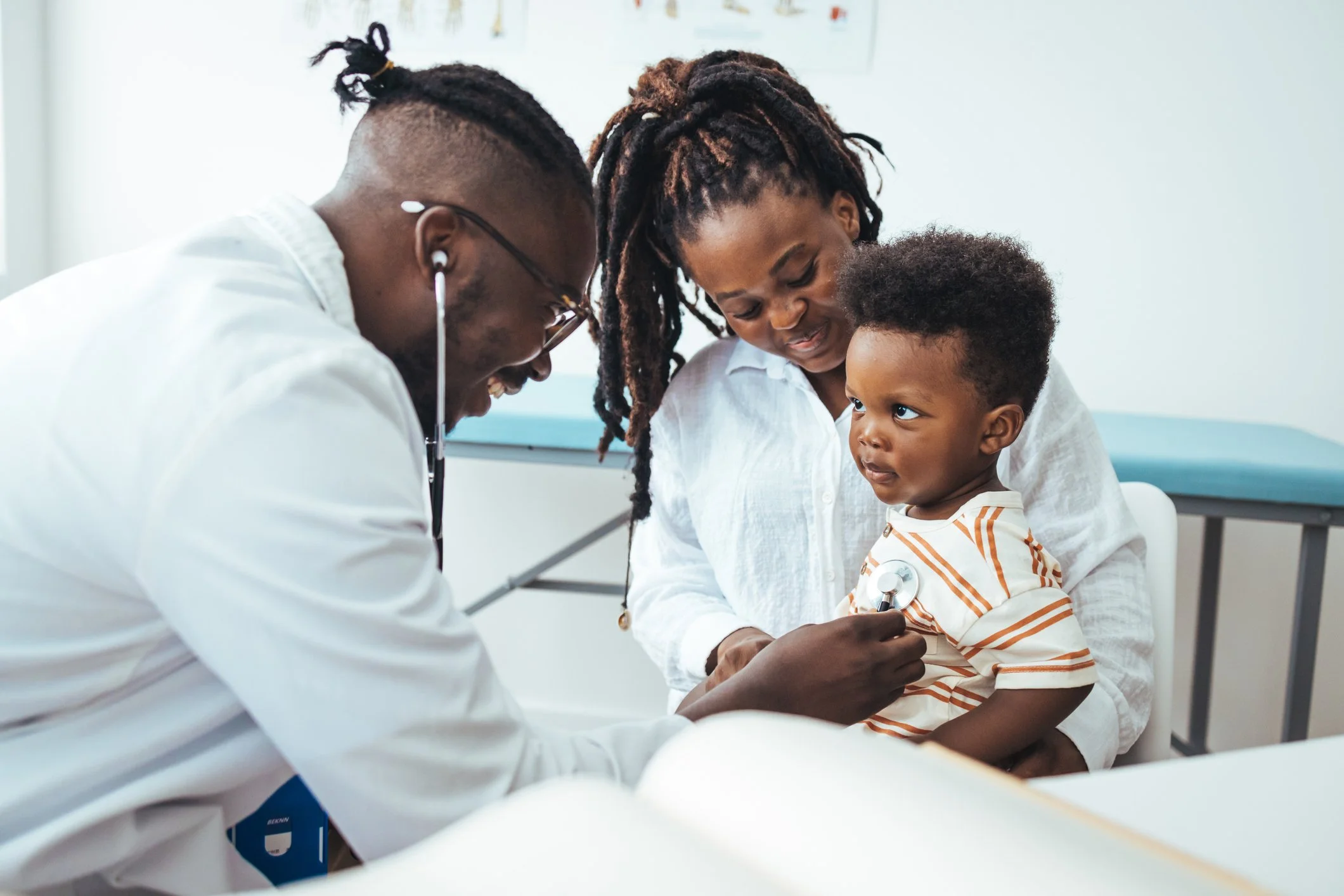A doctor using a stethoscope on a young boy sitting on his mother's lap in a medical examination room.