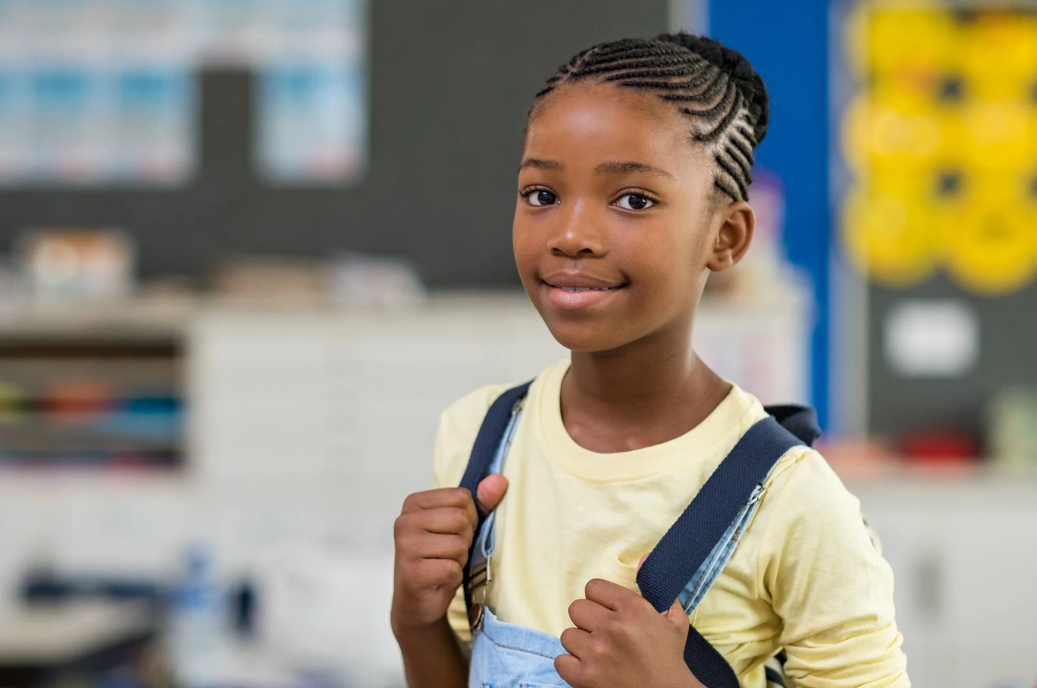 A young girl with braided hair and a yellow shirt, carrying a backpack, standing in a classroom or school setting.