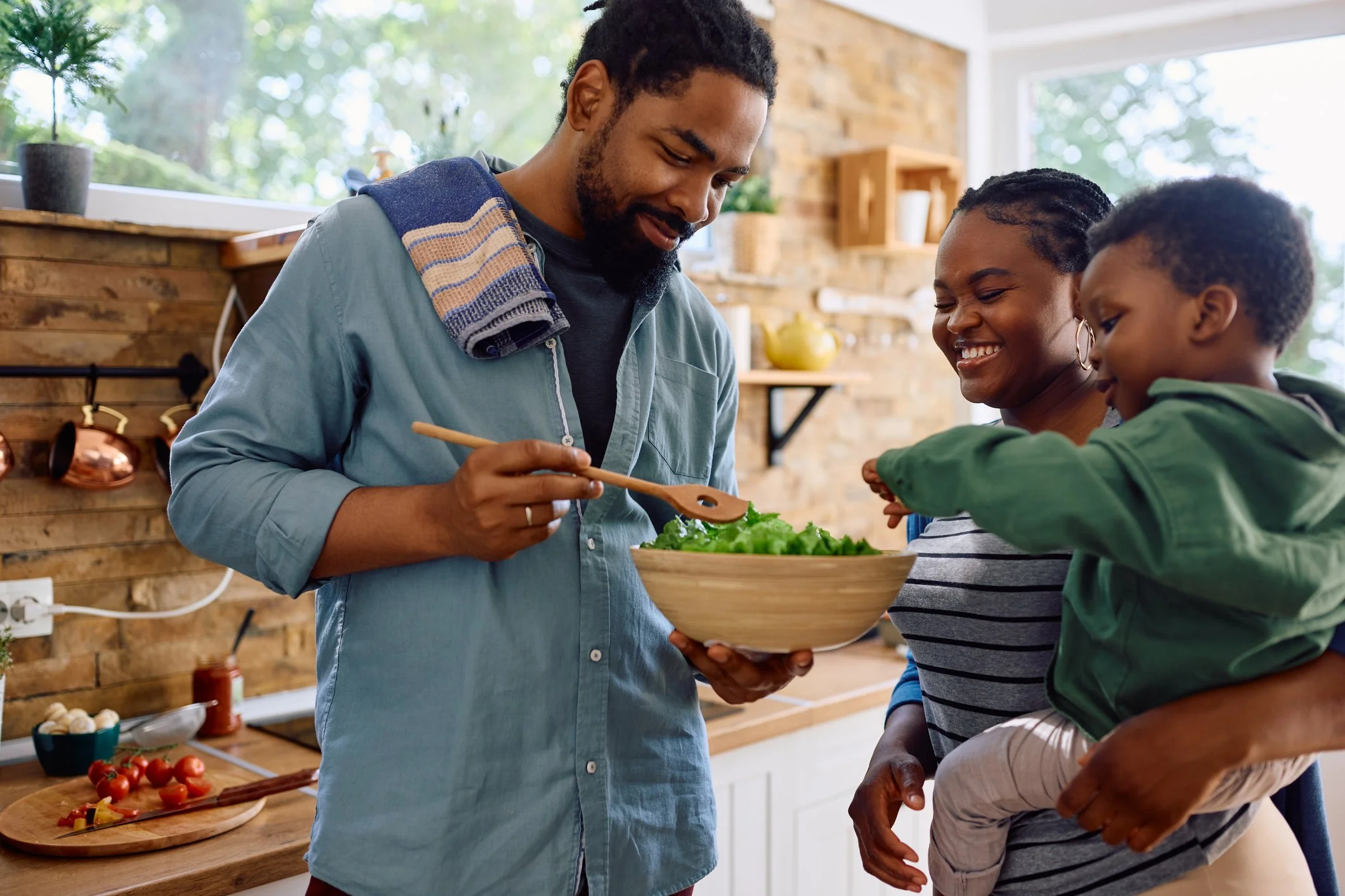 A family enjoying preparing a salad in a modern kitchen with wooden accents, sunlight coming through large windows.