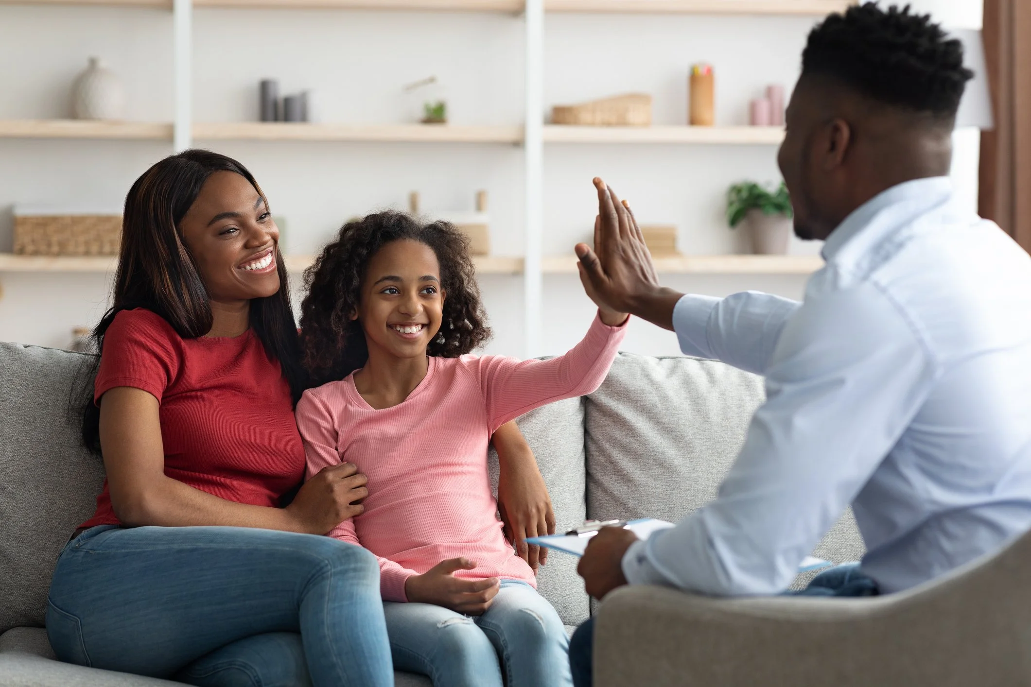 A mother, daughter, and a therapist are sitting on a sofa, smiling and giving each other high fives in a therapy or counseling session.