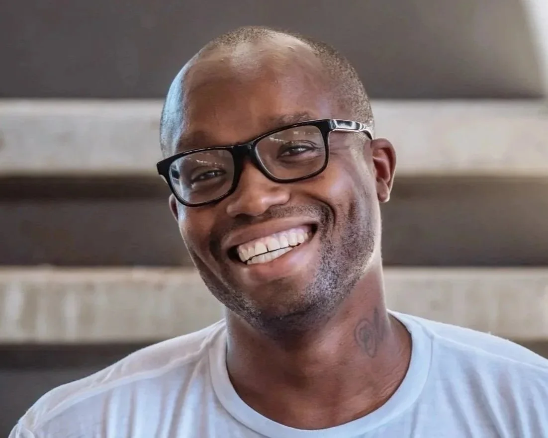 A smiling man with glasses, short hair, and a tattoo on his neck.