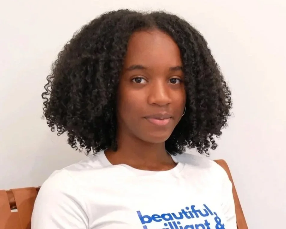 A young woman with curly black hair, wearing a white t-shirt with blue text, sitting on a wooden chair against a plain white wall.