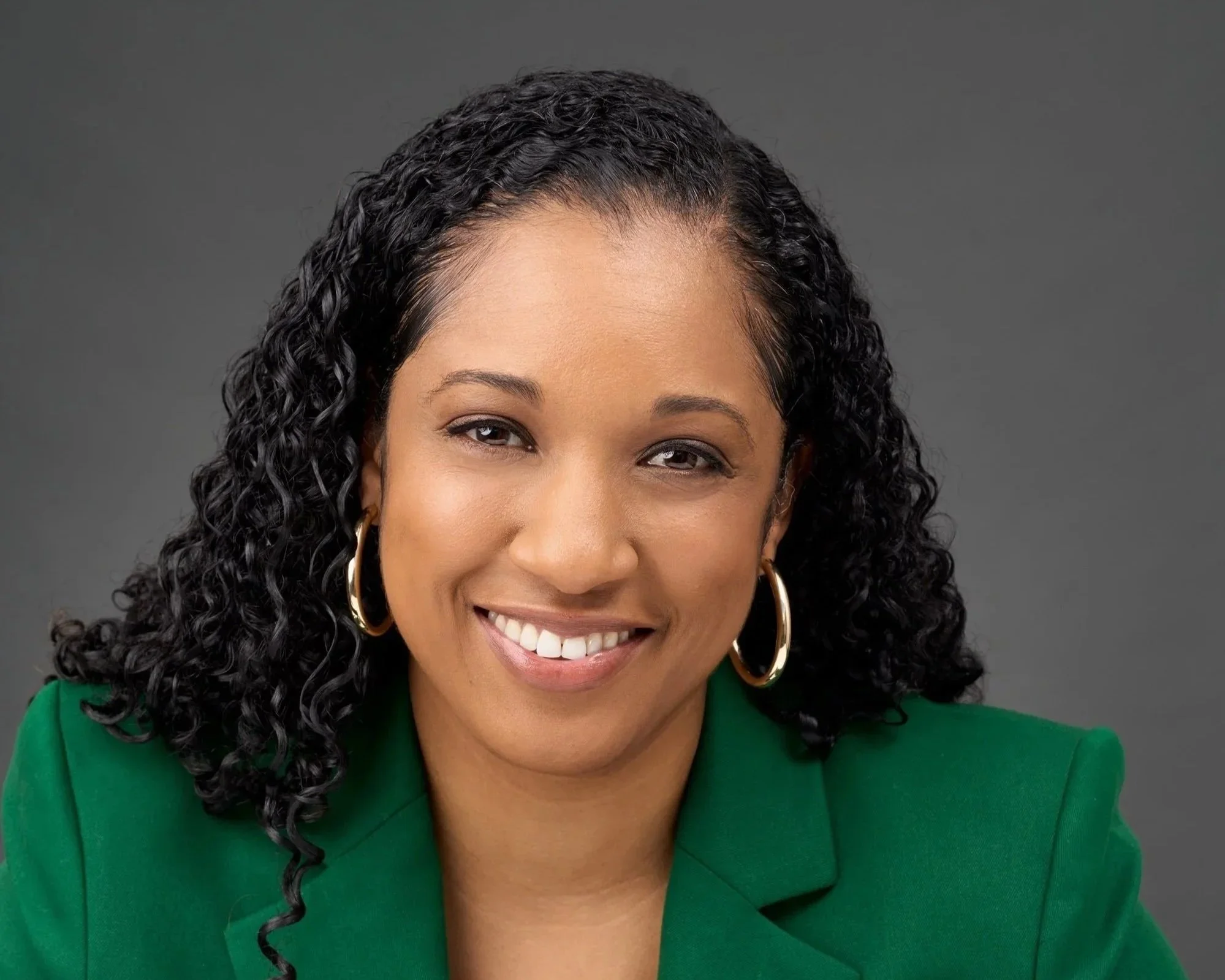 A woman with curly black hair wearing a green blazer and gold hoop earrings, smiling at the camera against a gray background.