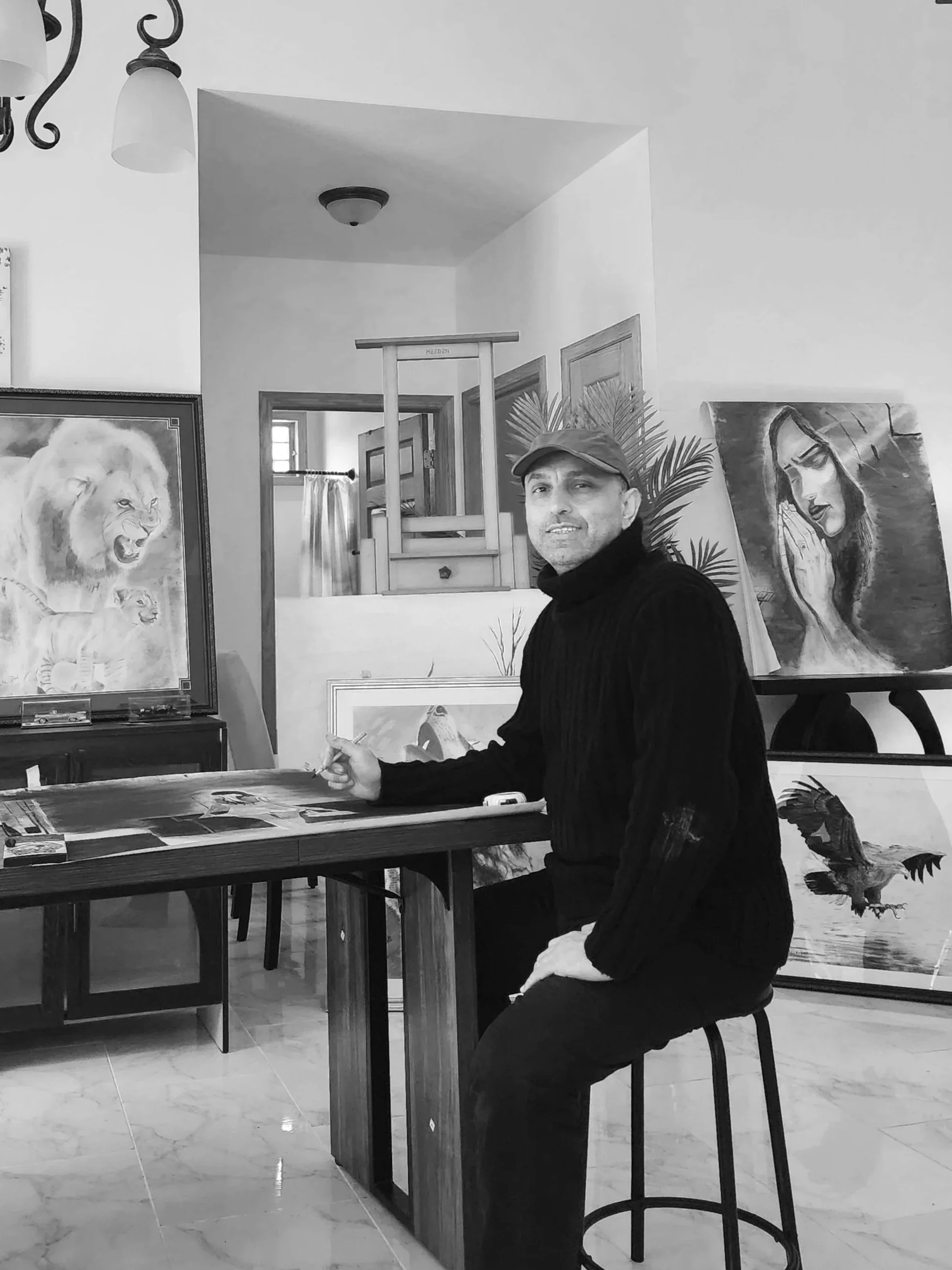 A man wearing a cap and dark sweater sitting on a high stool at a table in an art studio, with paintings of a lion, a woman praying, a fish, and a bird displayed in the background.