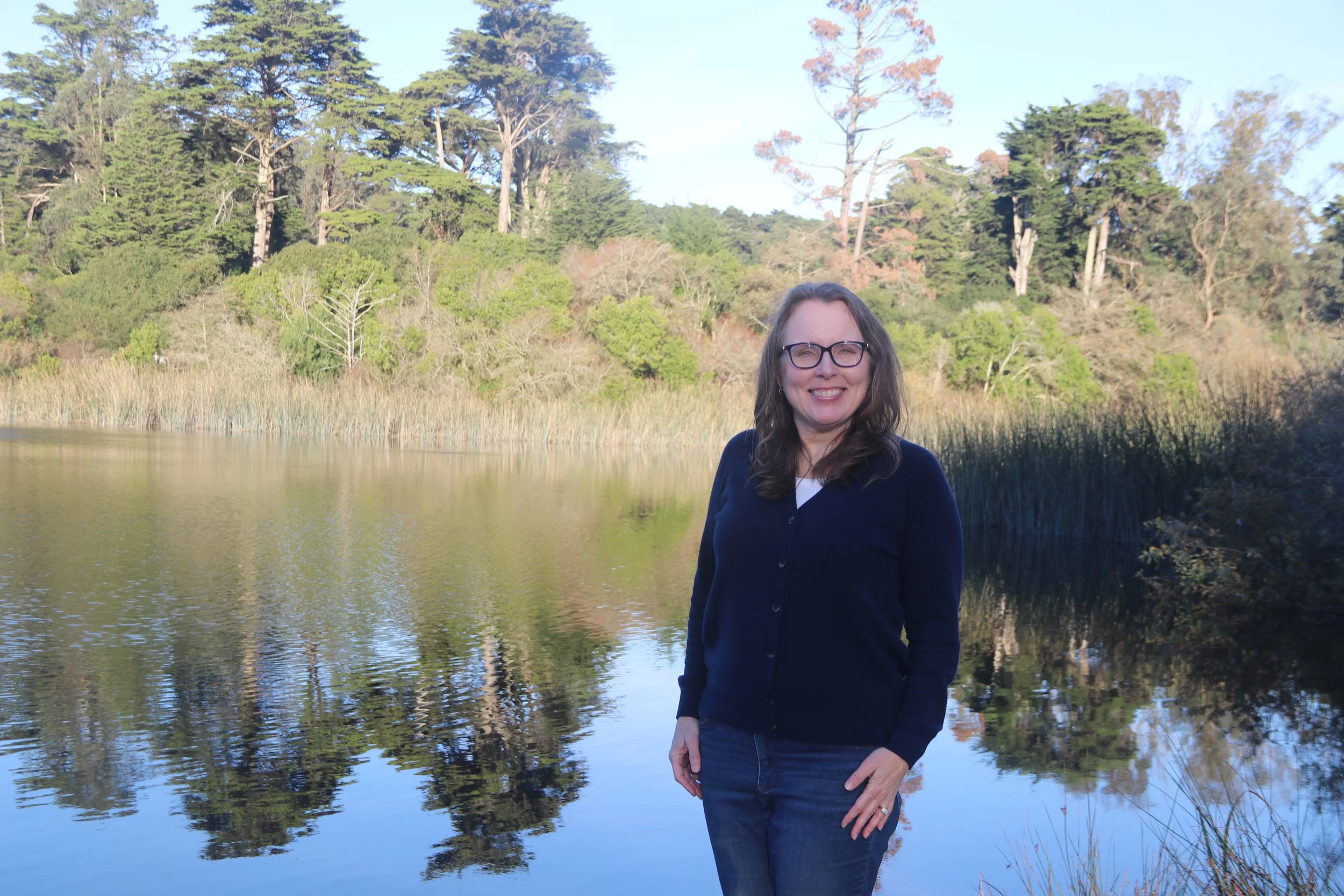 A woman with glasses wearing a navy blue sweater standing by a lake with trees and bushes in the background.