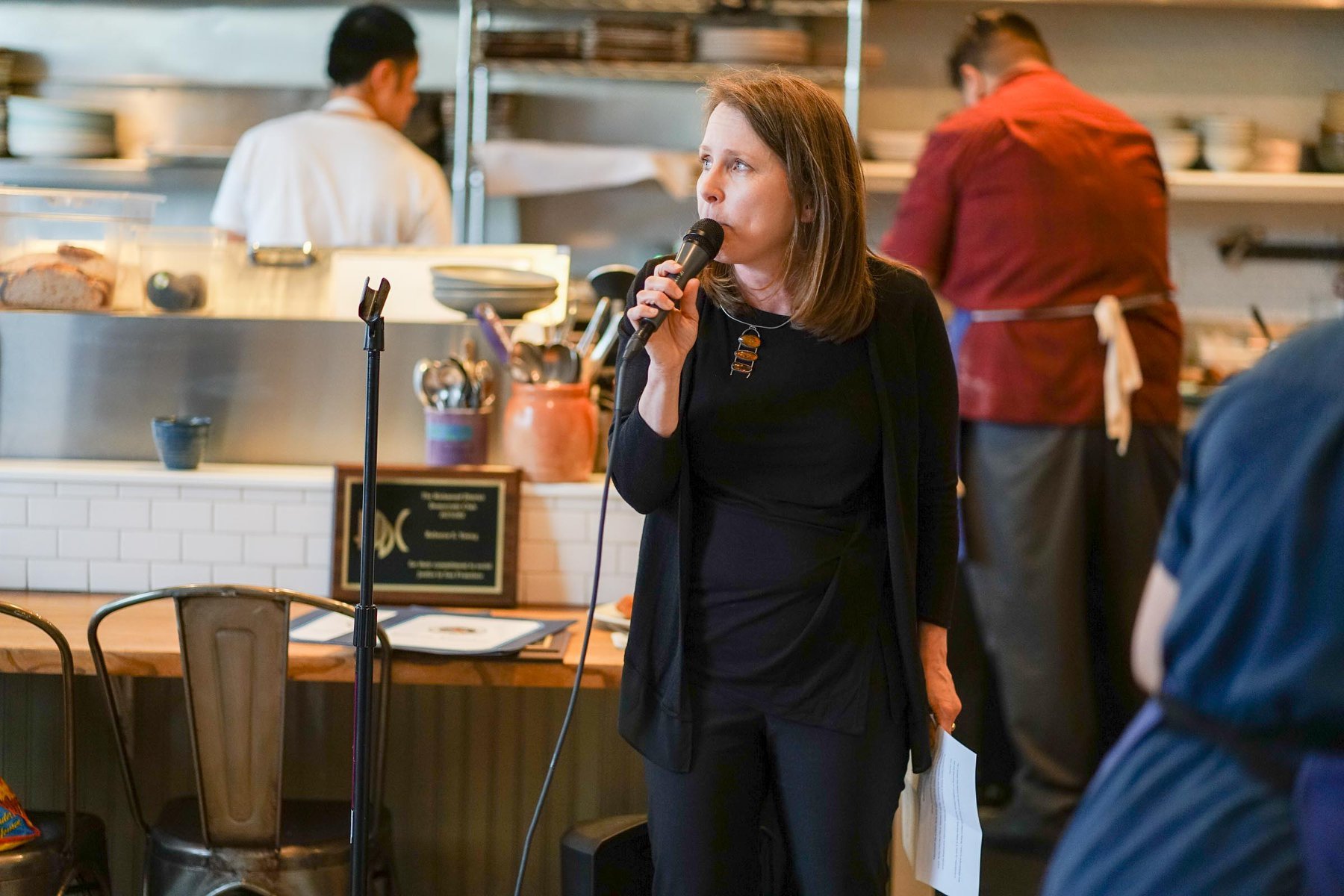 A woman standing and speaking into a microphone in a restaurant or cafe setting with kitchen staff working in the background.