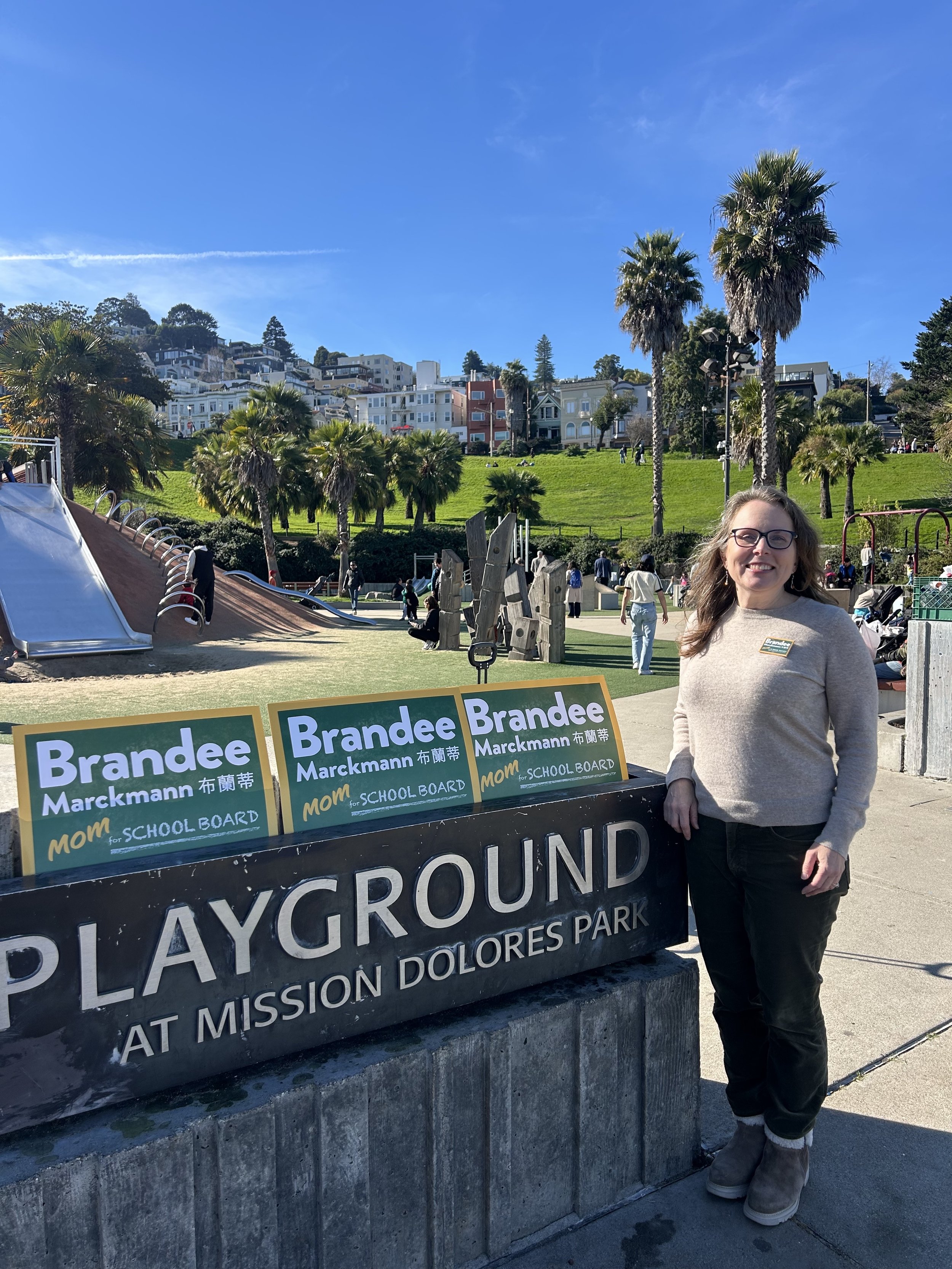 Woman smiling at the camera standing in front of a sign that says 'PLAYGROUND AT MISSION DOLORES PARK' with a playground and hillside houses in the background under a clear blue sky.