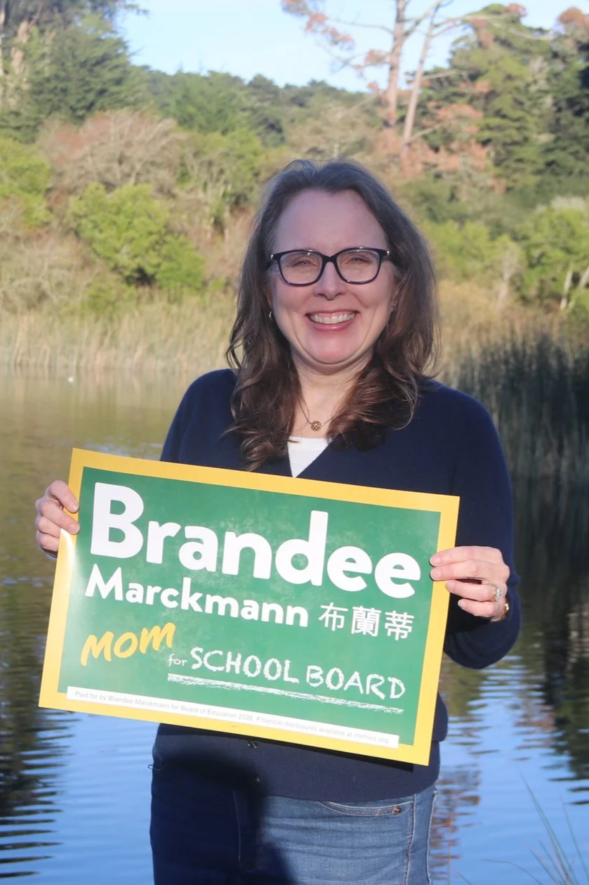 A woman with glasses smiling outdoors in front of a lake and trees, holding a campaign sign that reads 'Brandee Marckmann mom for SCHOOL BOARD'.