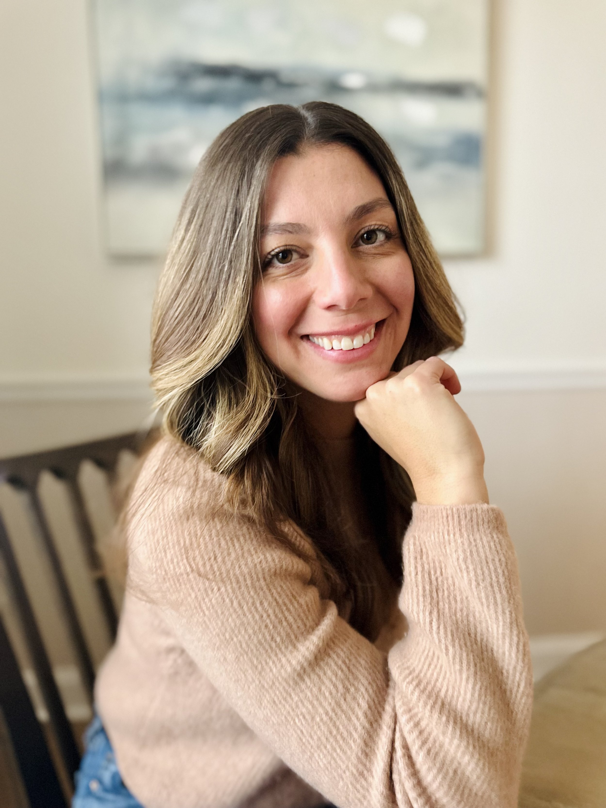 A woman, eating disorder therapist with long wavy brown hair smiling, resting her chin on her hand, sitting indoors with a blurred painting in the background.