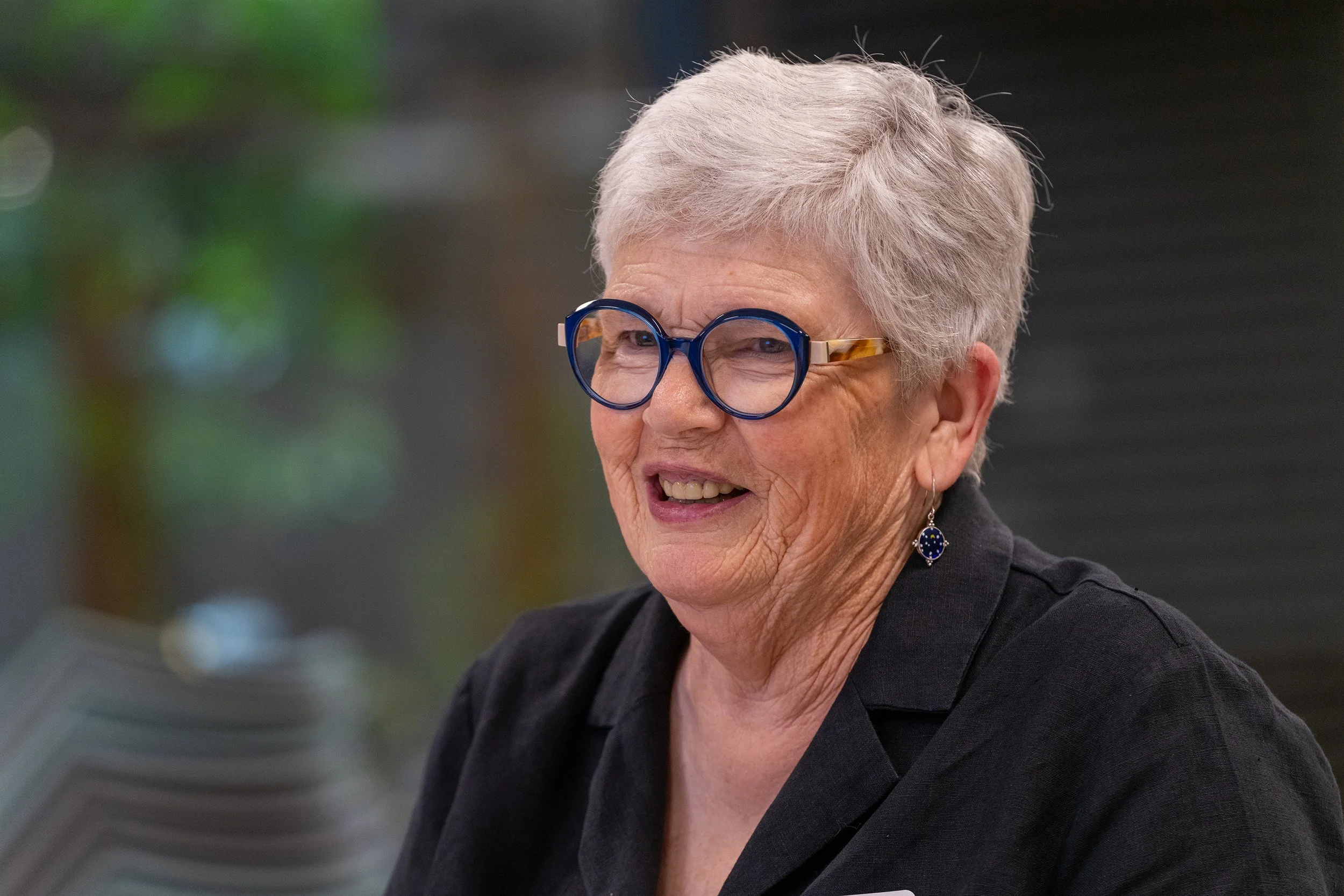 A smiling older woman with short gray hair wearing large round blue glasses, earrings, and a black top, sitting in a well-lit indoor space.