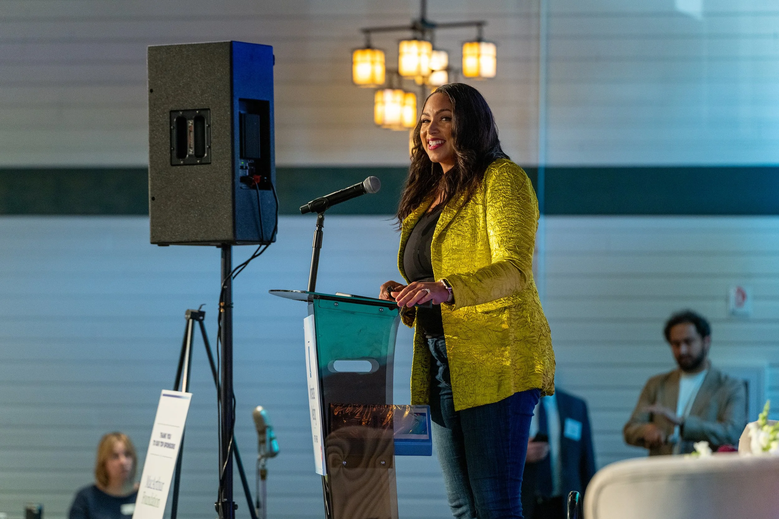 A woman stands at a podium with a microphone, smiling, wearing a yellow jacket, during a conference or event. There are people seated in the background.