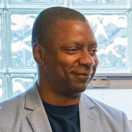 Man in a gray blazer smiling, standing indoors near a glass block wall.