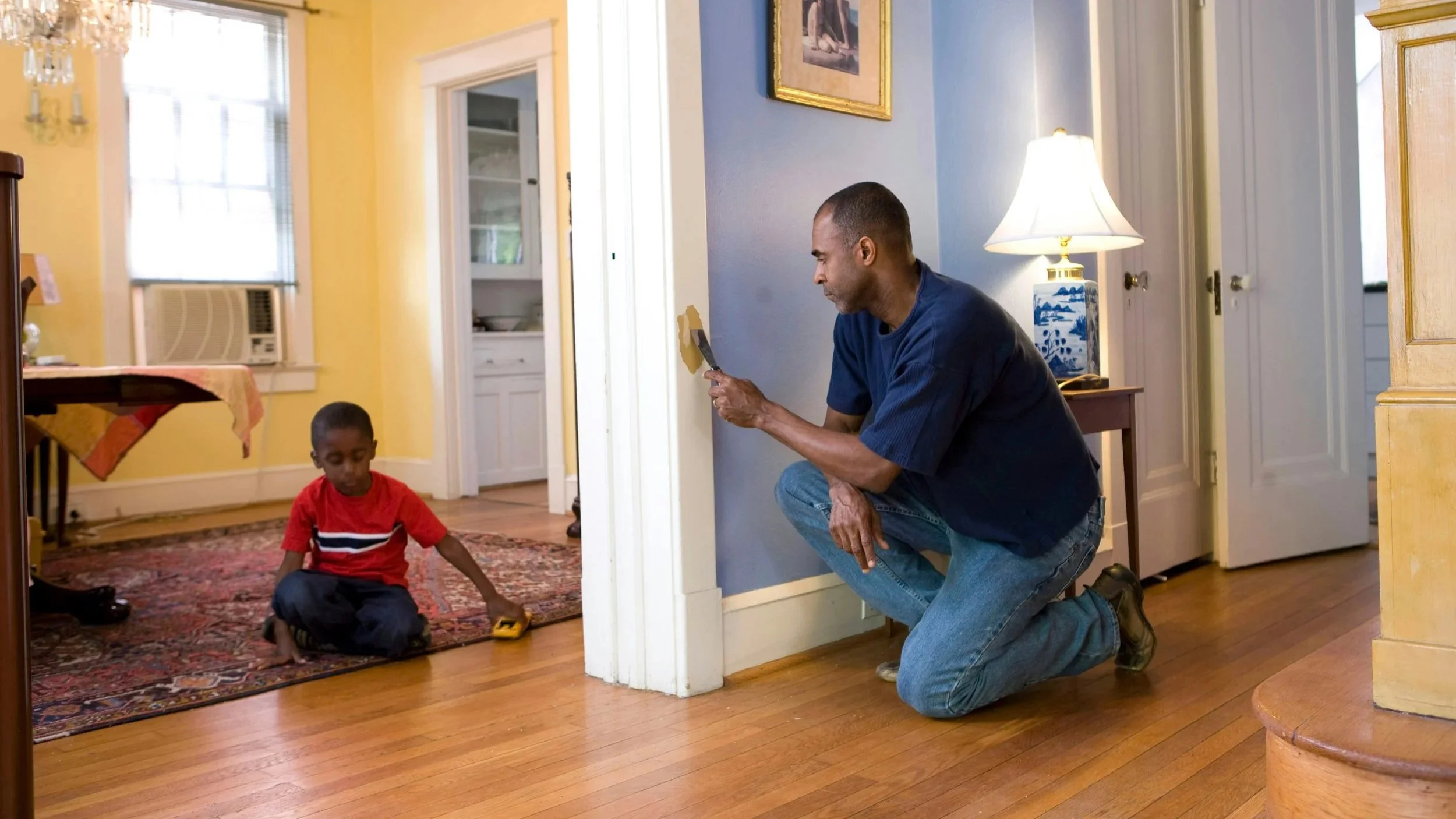A man kneeling on the floor and a young boy sitting on a rug inside a house. The man is holding a paint spatula and appears to be fixing a wall. The boy is playing with a small toy car.