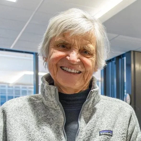 An elderly woman with short gray hair smiling, wearing a gray Patagonia jacket and a black shirt inside a building with large windows.