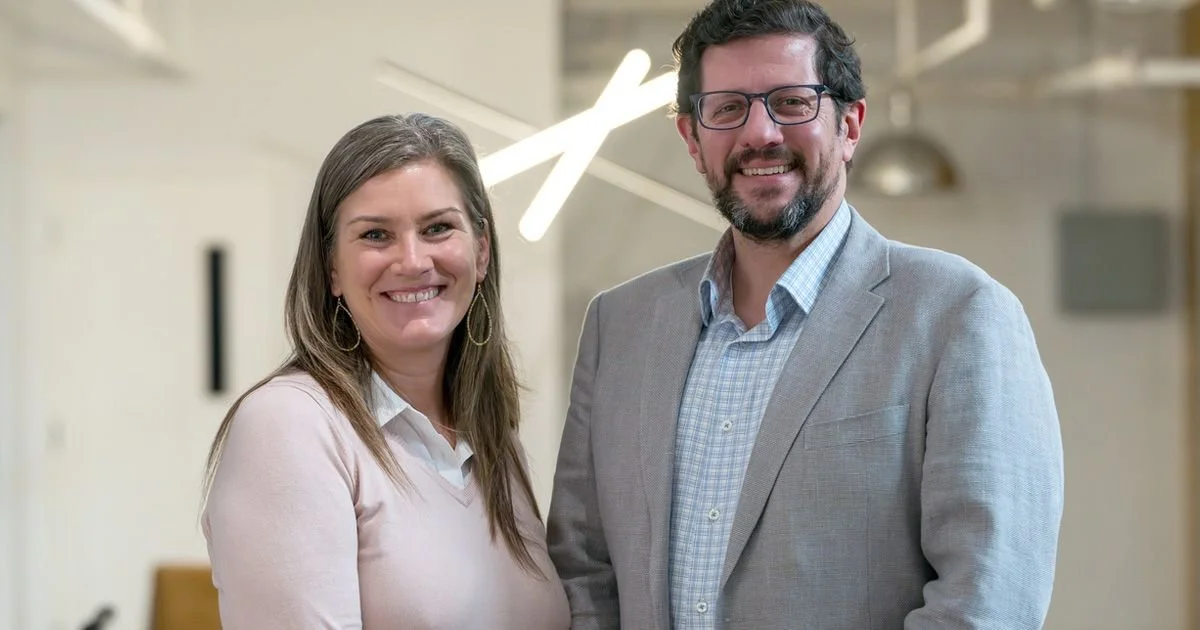 Two people in business attire standing in an office, smiling at the camera.