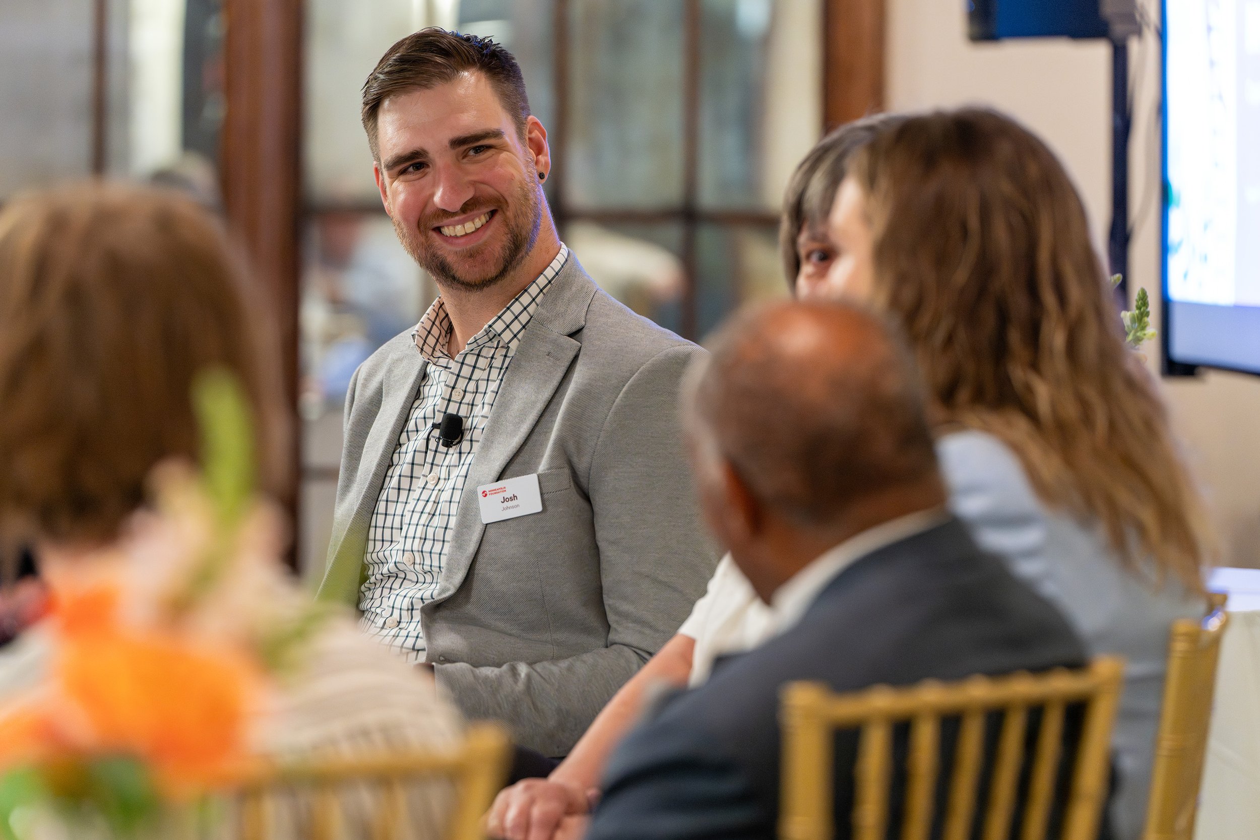 A man in a light gray blazer and checkered shirt smiling at a group meeting or panel discussion, with other people seated around a table.