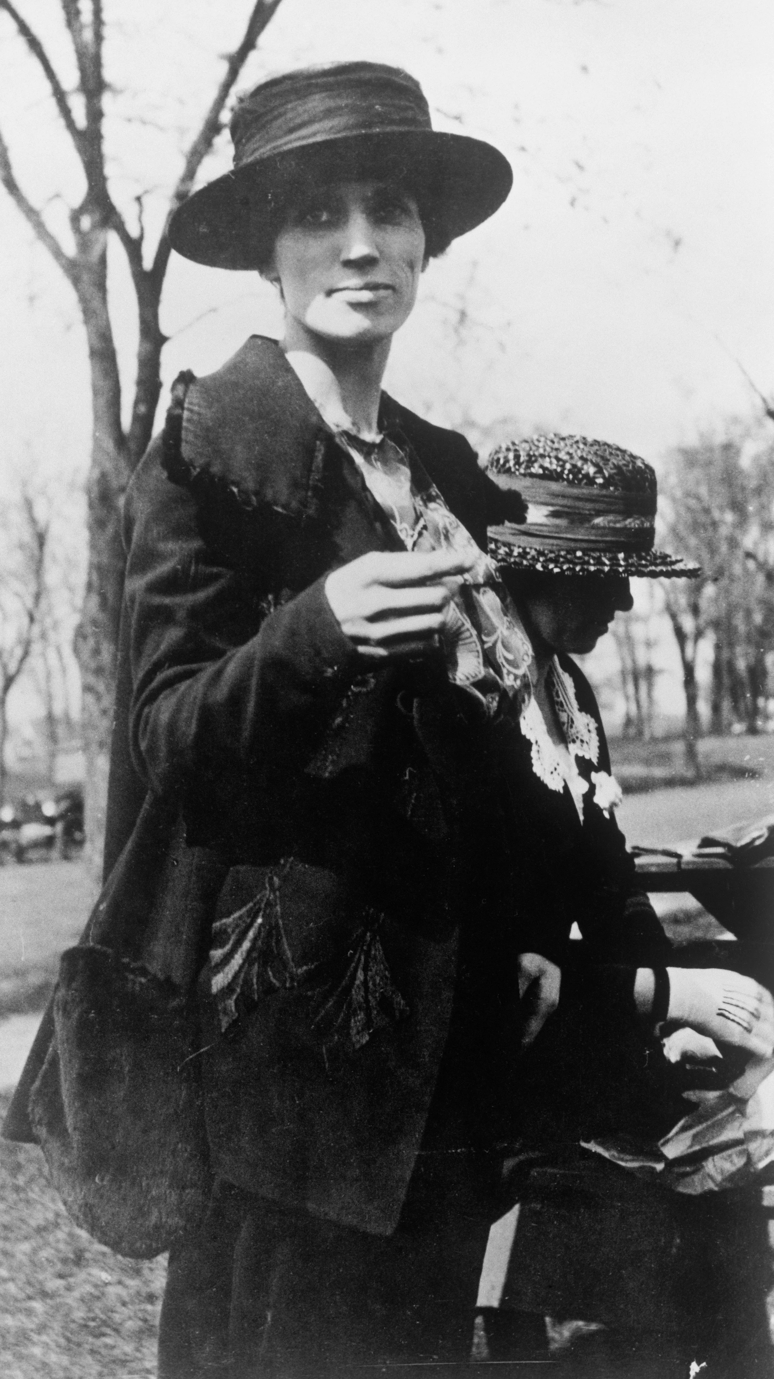 Black-and-white photo of a woman with light skin wearing a wide-brimmed hat, holding a cigarette, and standing outdoors. She has short hair and is dressed in a dark jacket and patterned blouse. A person with darker skin and a patterned hat is seated beside her, partially visible.