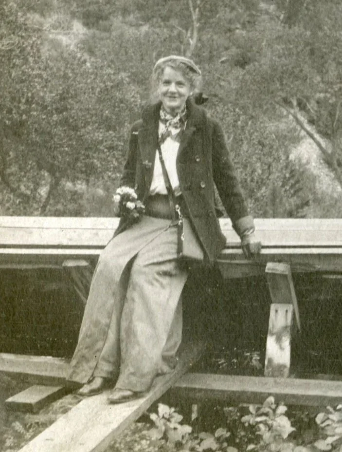 A black and white photo of a woman sitting on a wooden bridge in a natural outdoor setting, smiling at the camera. She is dressed in vintage clothing, including a long skirt, a jacket, and a scarf. She is holding a small bouquet of flowers in one hand and has gloves on. The background contains trees and foliage.