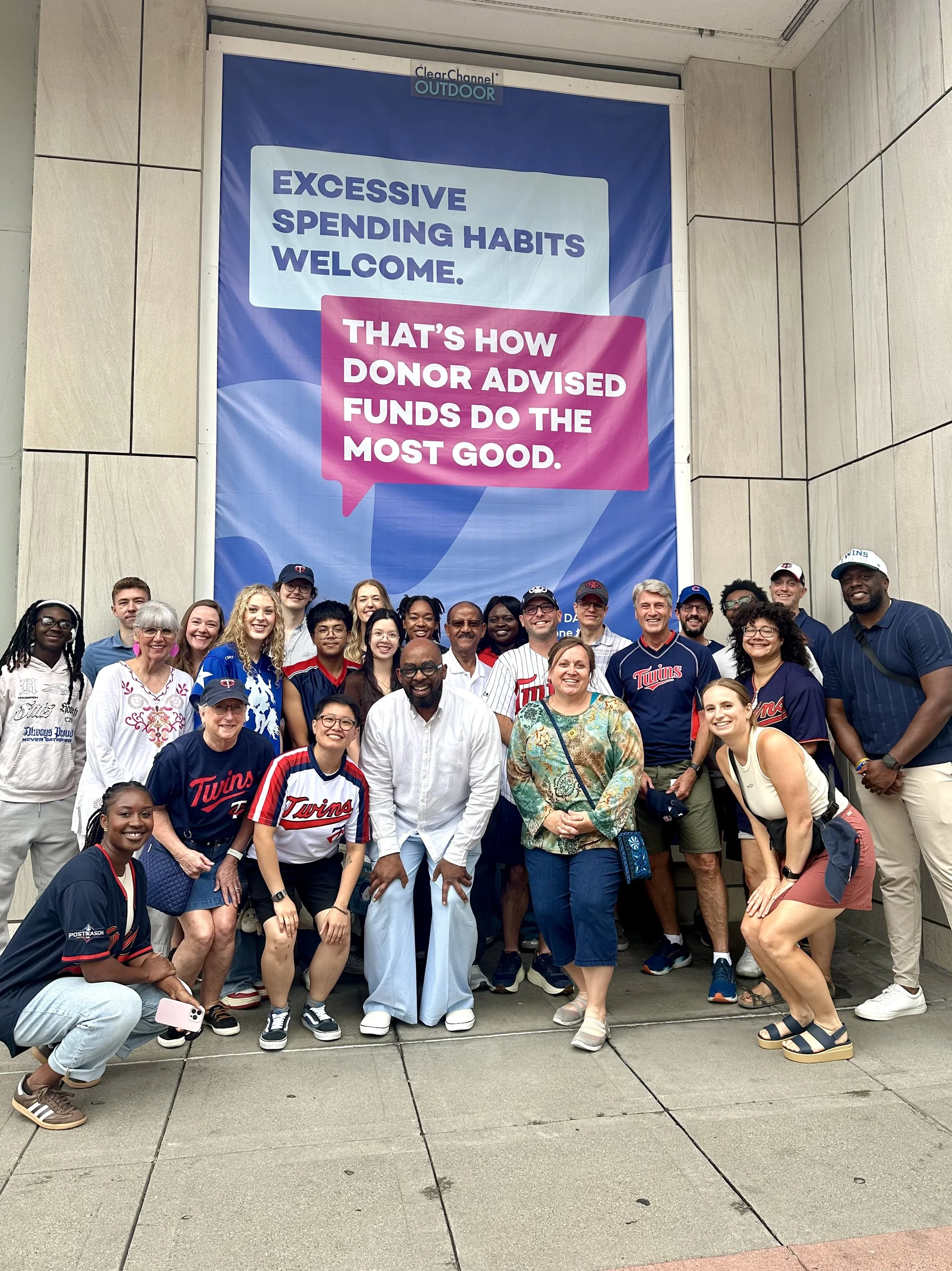 Group of people posing for a photo outside in front of a large sign with motivational messages, some wearing Minnesota Twins baseball jerseys and caps.
