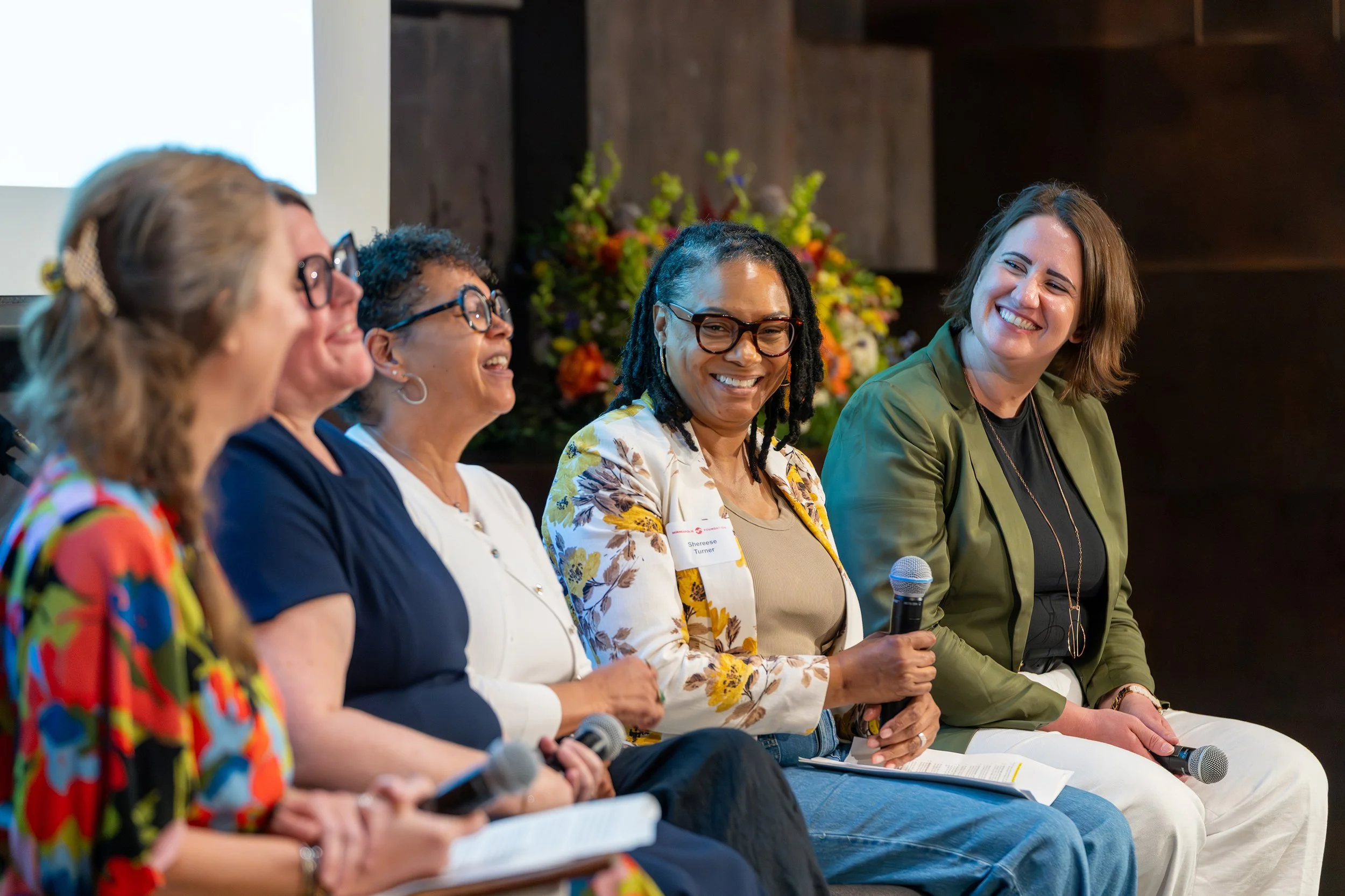 Five women sitting on a stage, participating in a panel discussion, smiling and holding microphones, with a flower arrangement in the background.