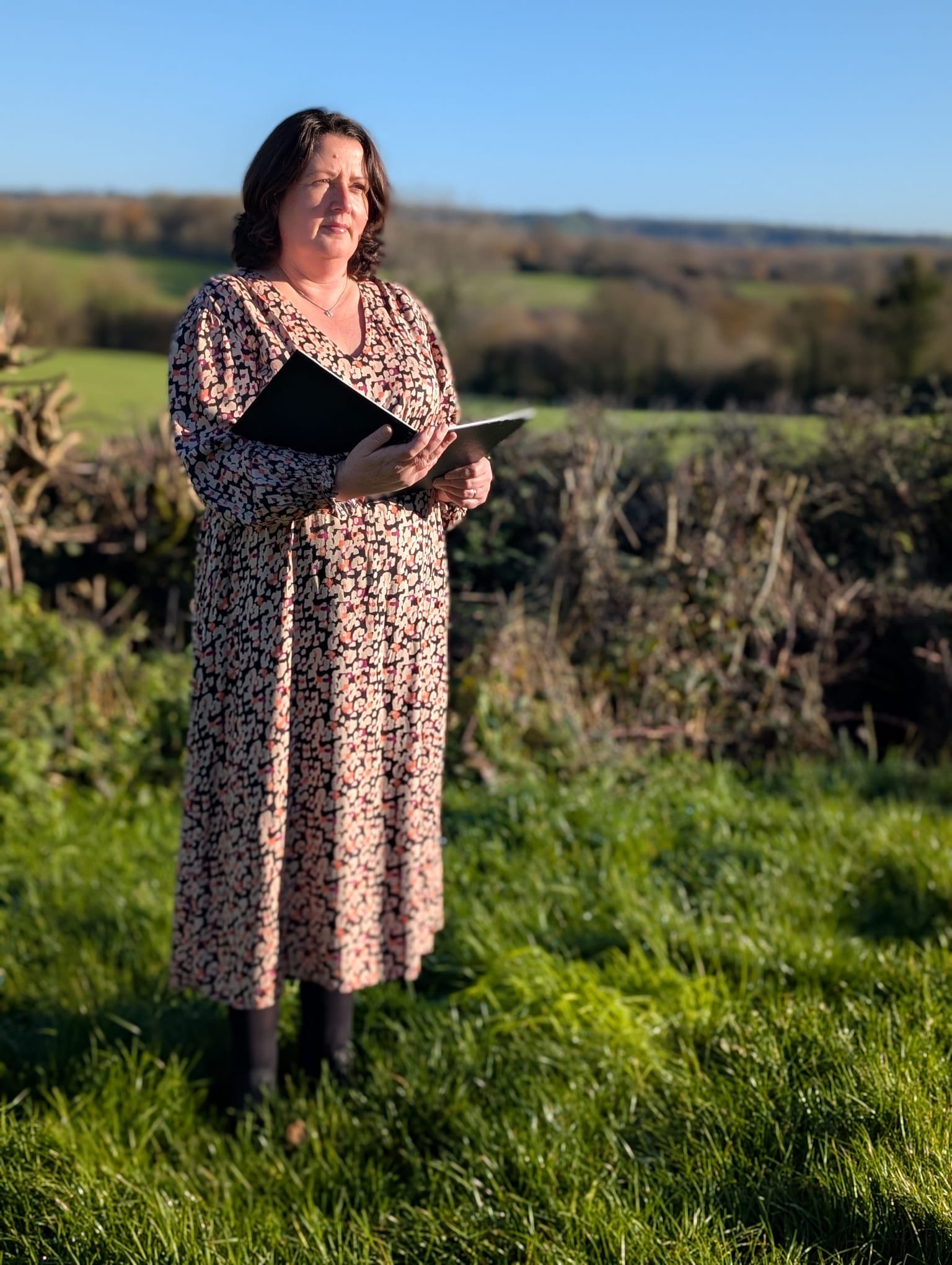 A woman standing outdoors on grass, holding a book or notebook, with a landscape of fields and trees in the background, under a clear blue sky.