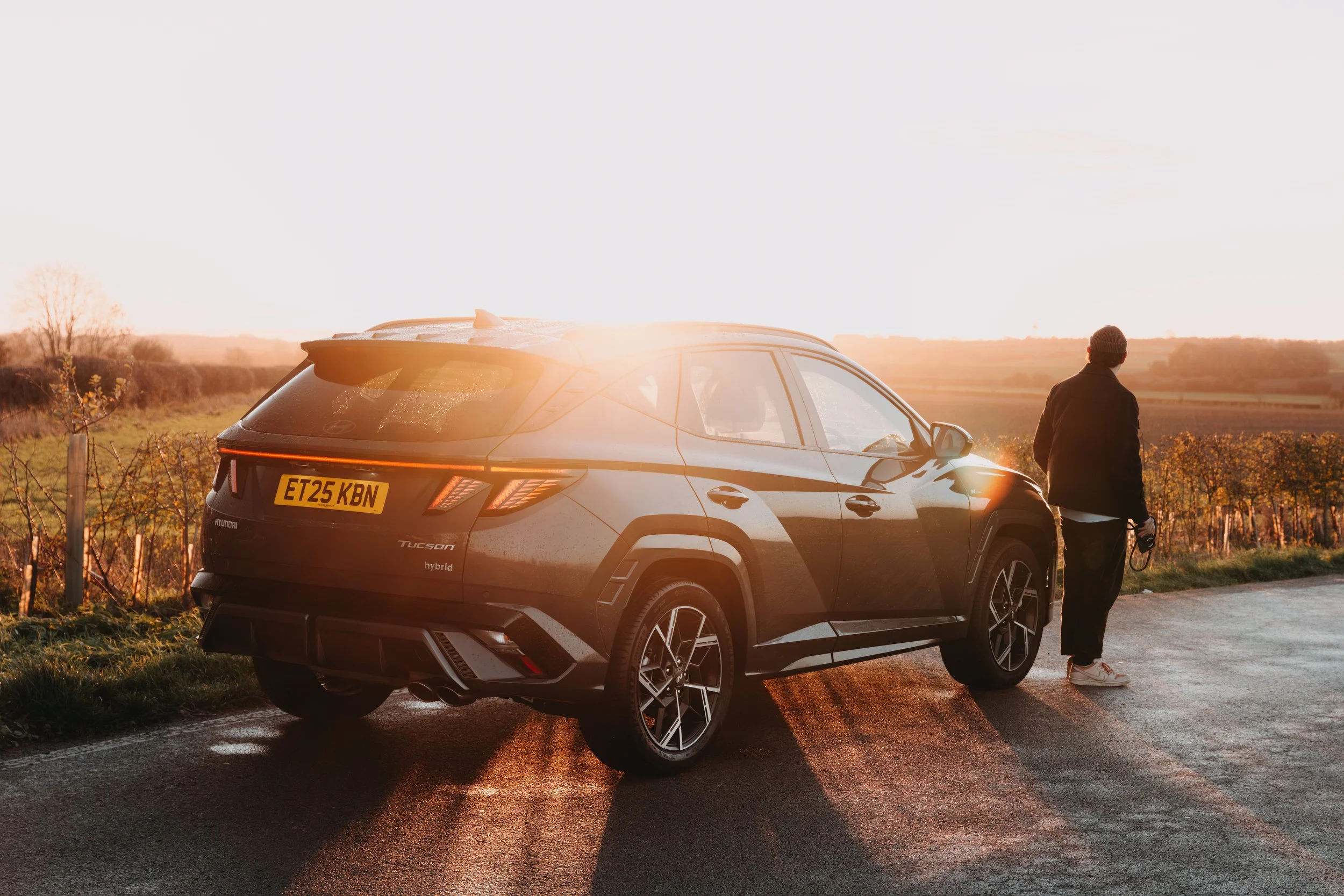 A person walking away from a black Hyundai Tucson hybrid SUV on a rural road during sunset, with fields in the background.