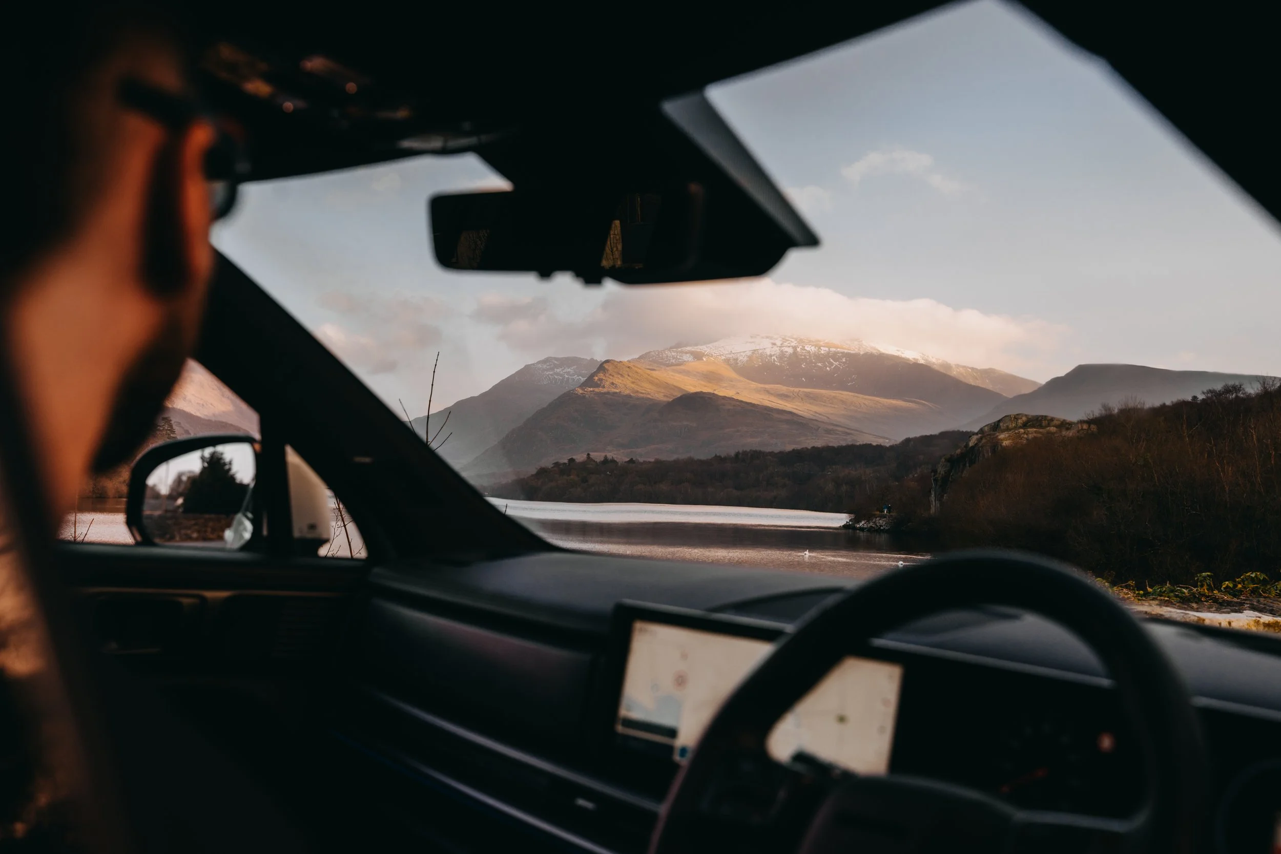 View from inside a car showing a driver looking out at a landscape with mountains, a body of water, and a partly cloudy sky.