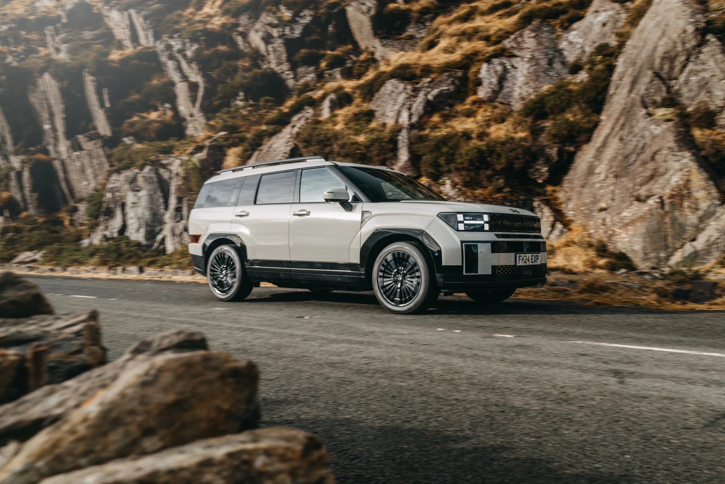 A futuristic white SUV driving on a mountain road with rocky cliffs in the background.