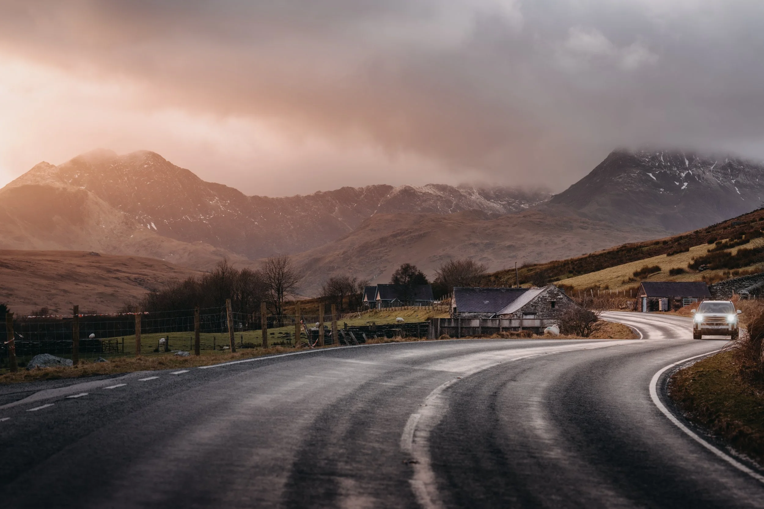Curving rural road leading towards houses with mountains in the background under a stormy sky.