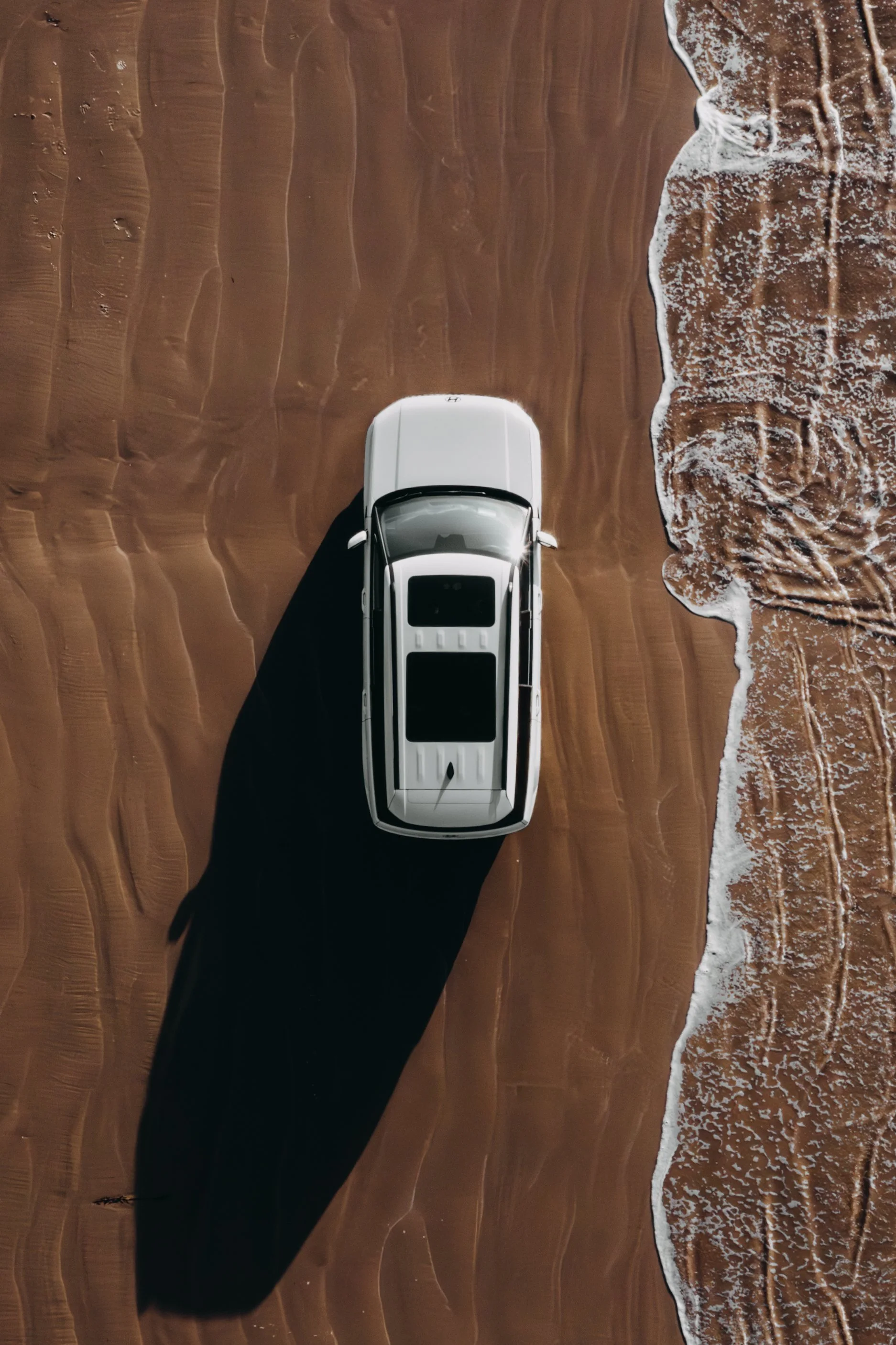 Top-down aerial view of a white SUV parked on a sandy beach with waves washing onto the shore.