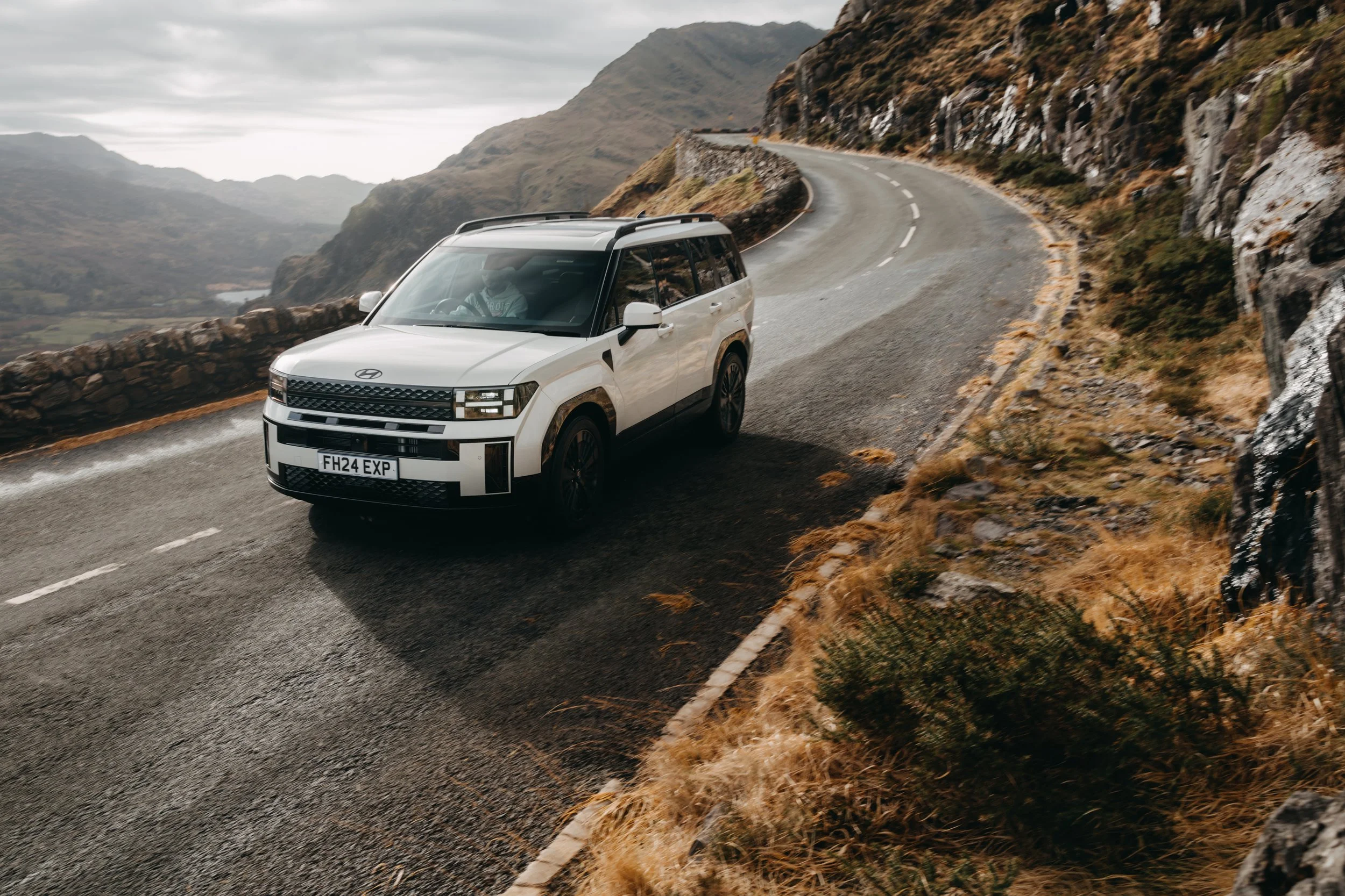 A white SUV driving on a winding mountain road with rocky cliffs and distant hills.