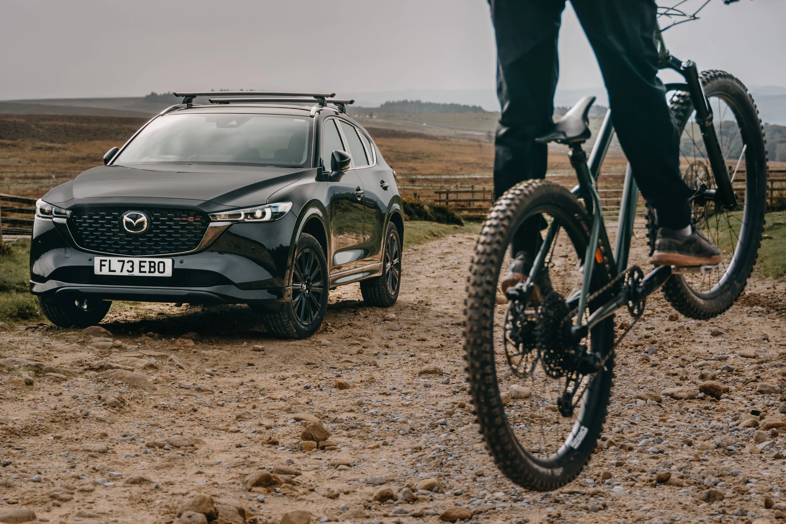 A black Mazda SUV on a dirt trail in a rural landscape with mountains in the background, and a person riding a mountain bike in the foreground.