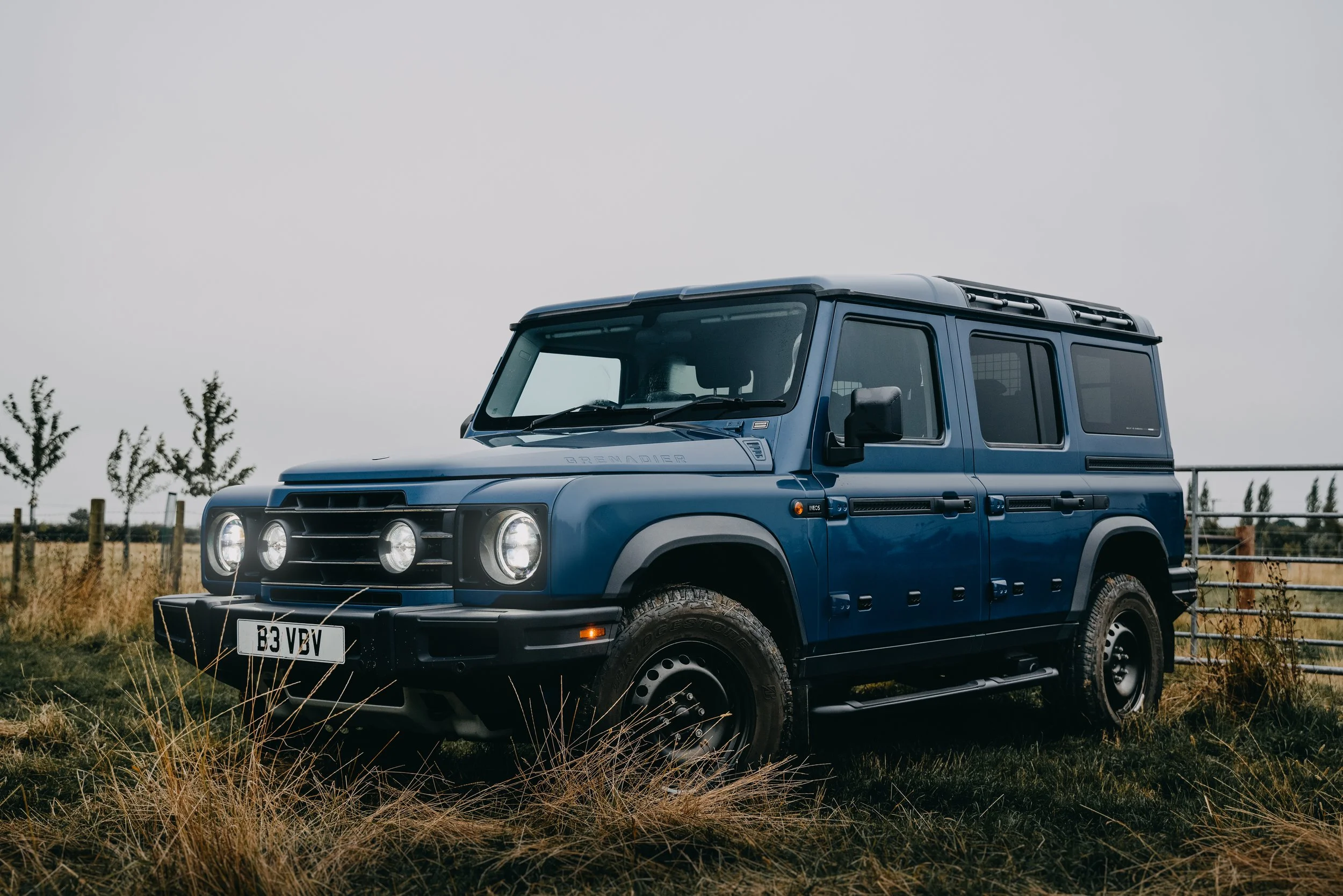 A blue Land Rover Defender vehicle parked on a grassy field with a fence and trees in the background under a cloudy sky.