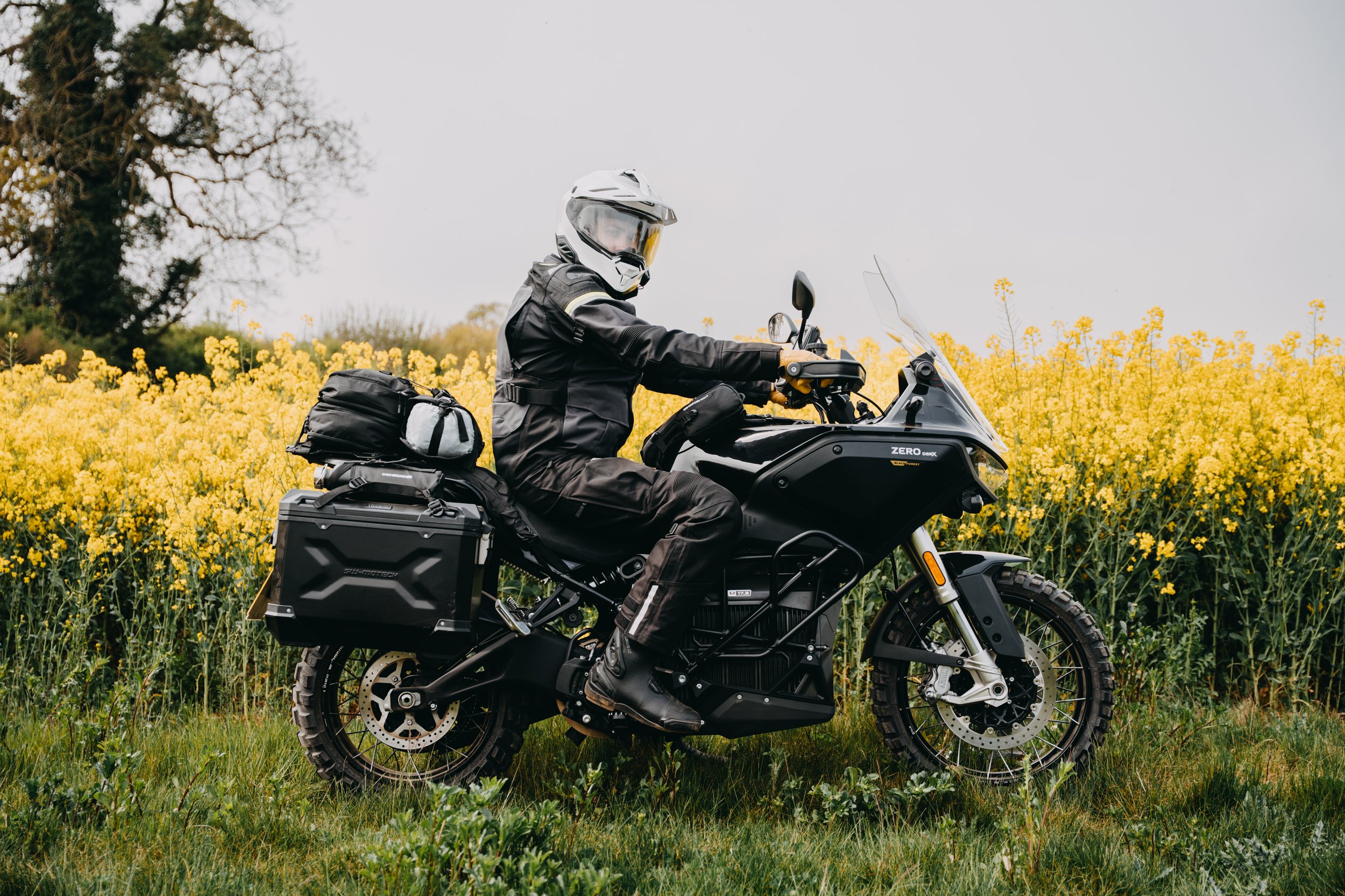 Motorcycle rider dressed in black gear and helmet riding through a field of yellow flowers with a cloudy sky in the background.