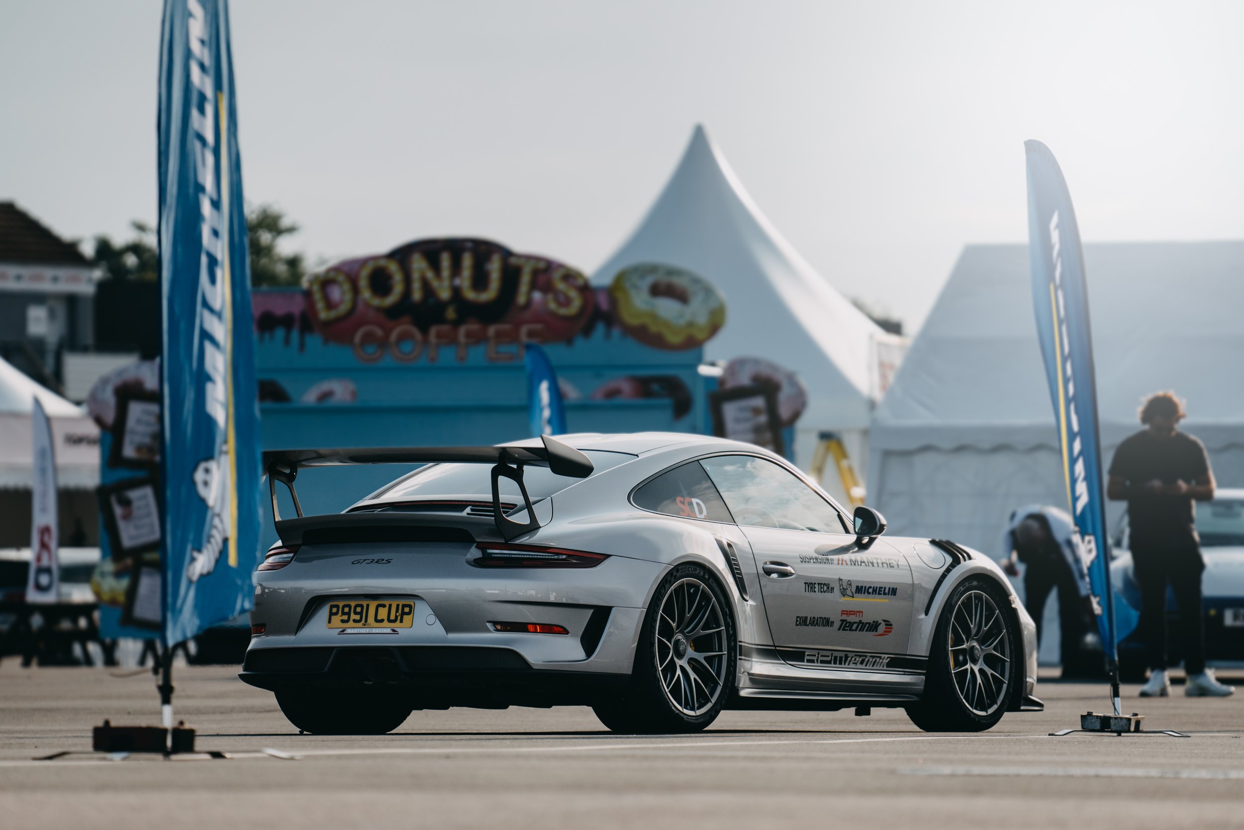 A silver Porsche 911 GT3 RS race car parked at a motorsport event with sponsorship stickers on the side, surrounded by blue motorsport banners and tents, with a donut stand in the background.