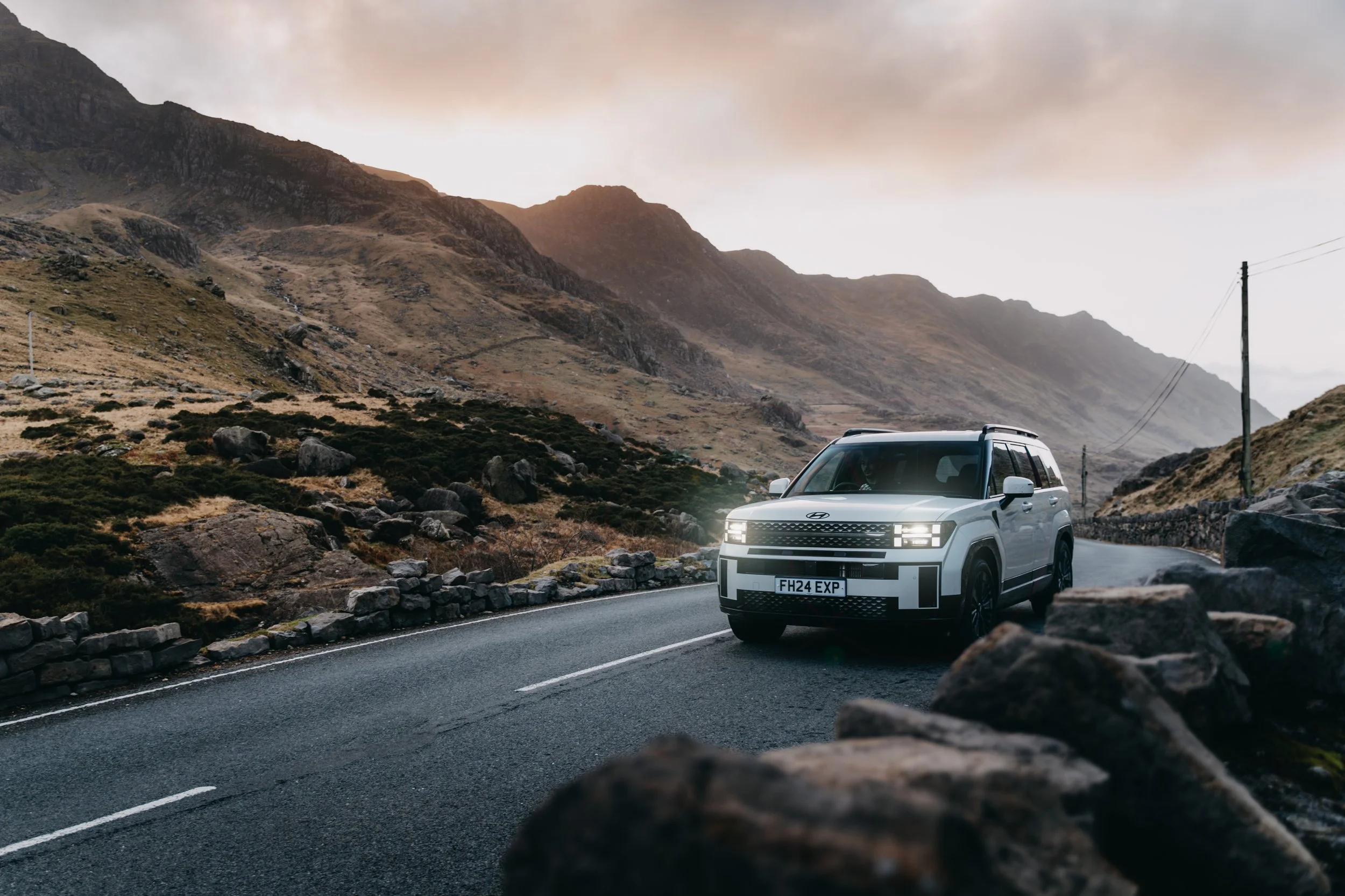 A white SUV driving on a mountain road with rocky terrain and hills in the background, cloudy sky overhead.