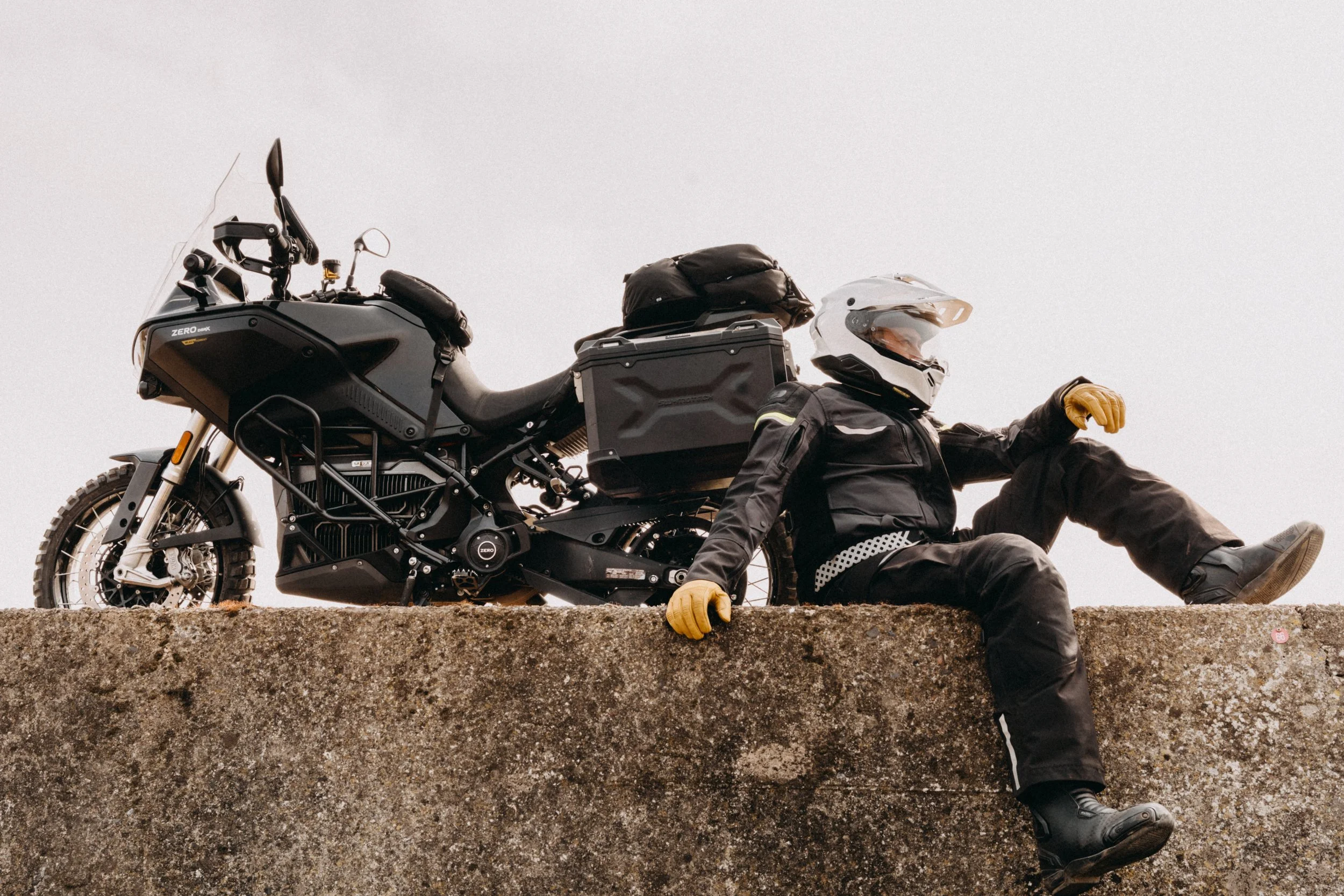 A person in motorcycle gear sitting on a concrete ledge next to a black adventure motorcycle, with a helmet on and bags on the bike.