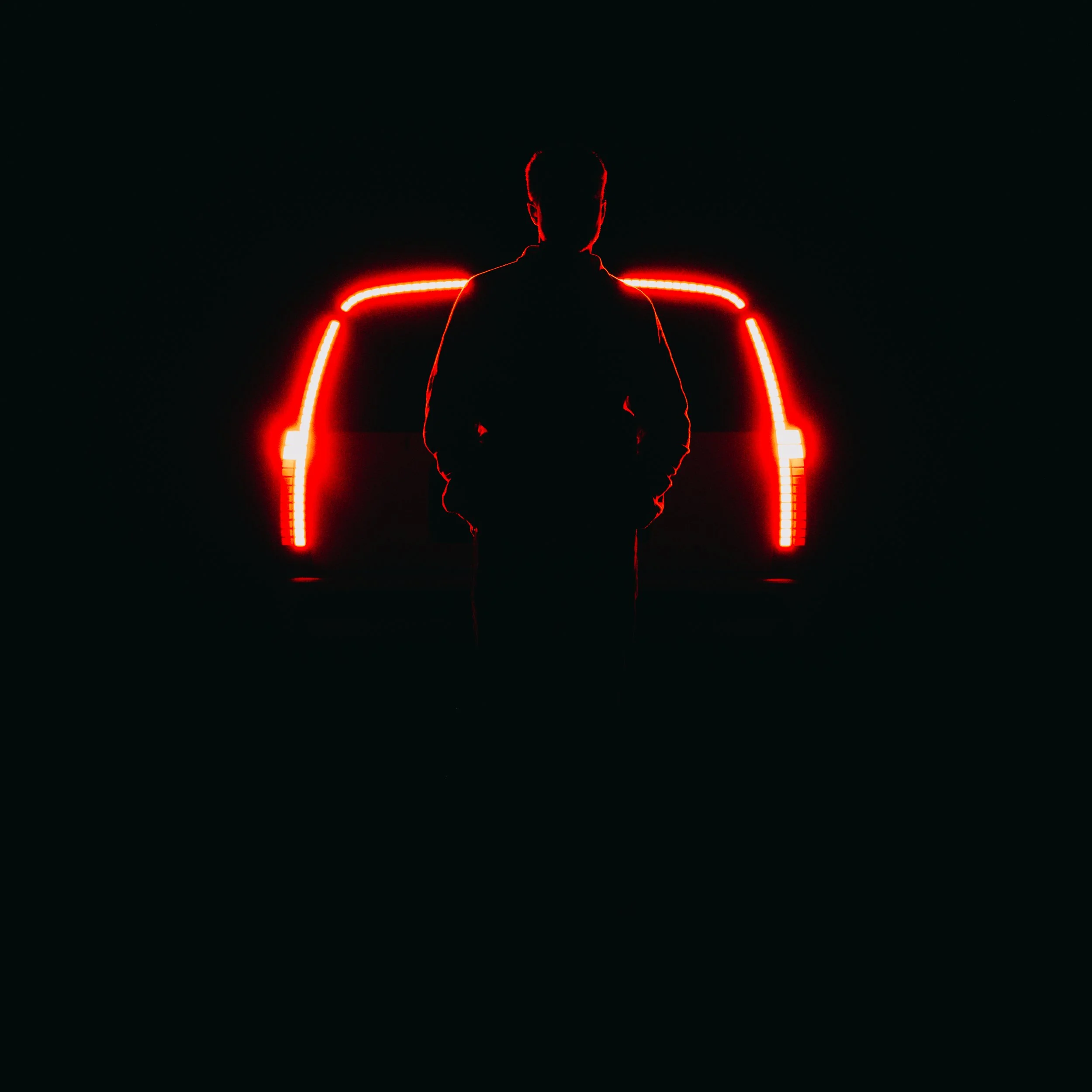Silhouette of a person standing in front of a vehicle with bright red rear lights at night.