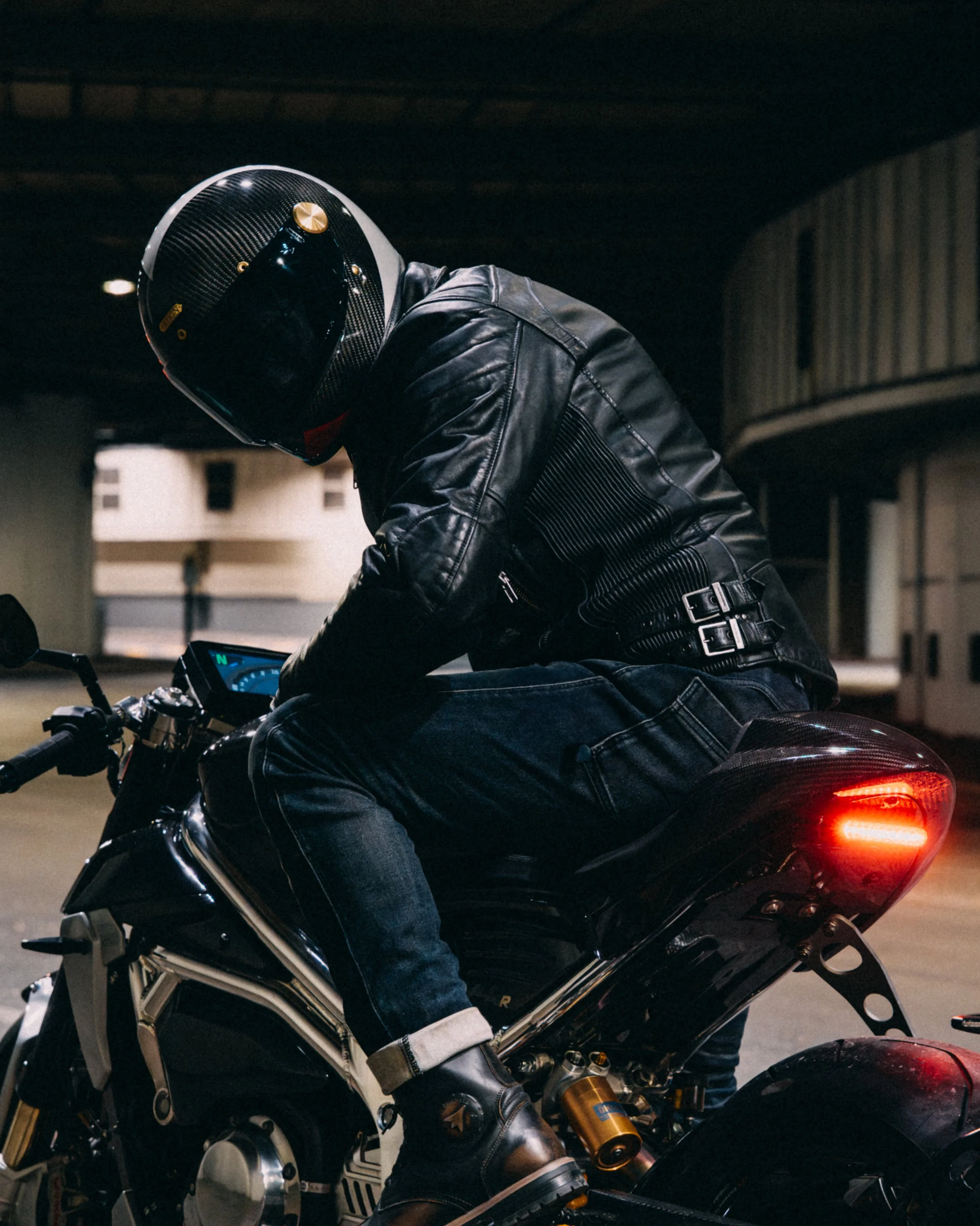 Motorcycle rider wearing a full face helmet and leather jacket, sitting on a motorcycle in an underground parking garage at night.