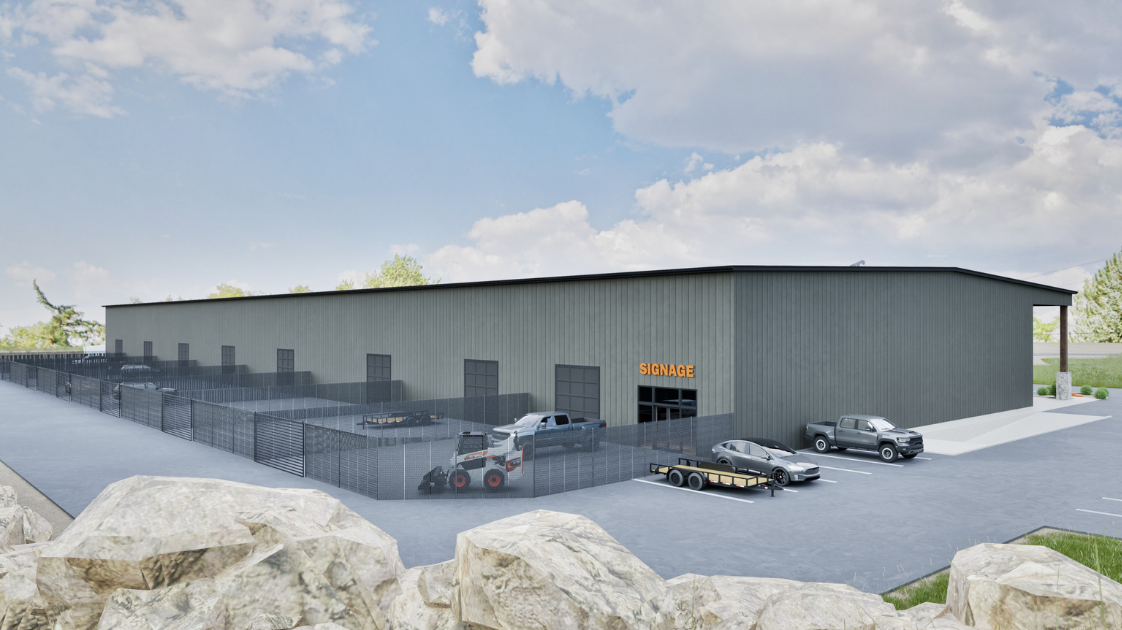 A large industrial warehouse with a fenced outdoor area containing parked vehicles including two trucks and a trailer, with a sign reading 'SIGNAGE' above the entrance, set against a partly cloudy sky.